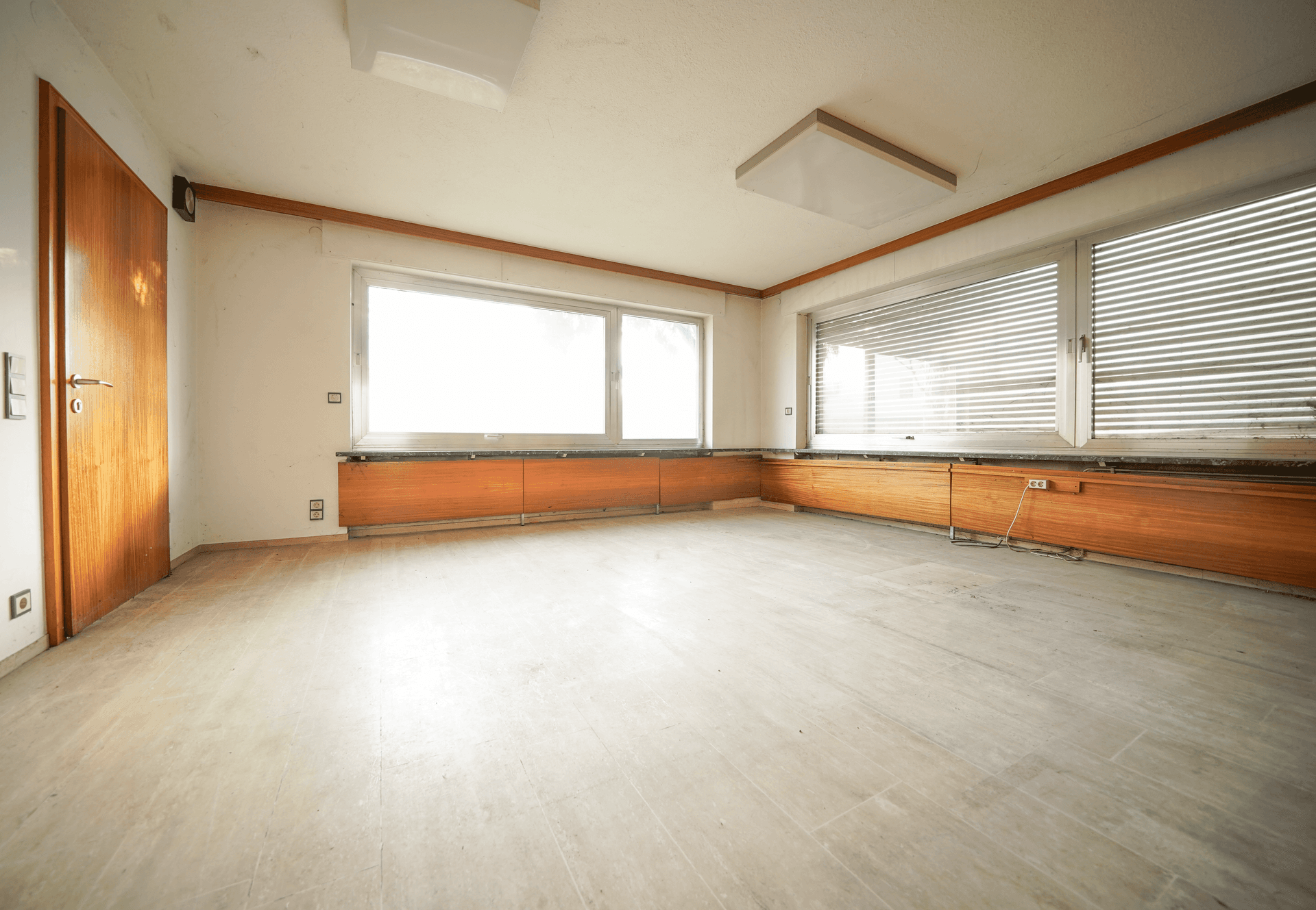 Geräumiges Zimmer mit hellen Fliesen, Fenstern mit Jalousien und Holzverkleidung unter den Fenstern.