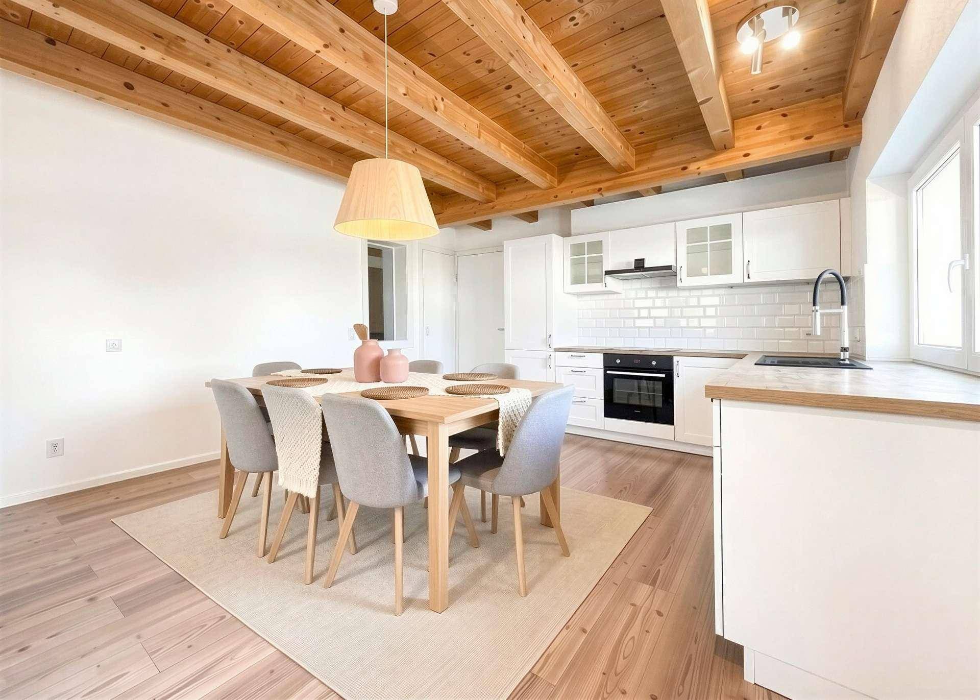 Heller Essbereich mit Holztisch und offener Küche unter einer Holzbalkendecke.