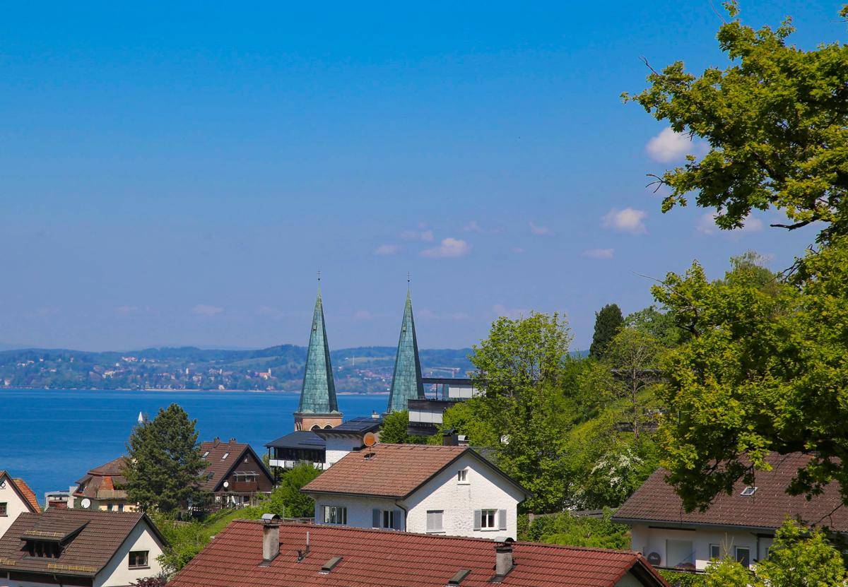 Blick über die Dächer auf zwei Kirchtürme und den weiten Bodensee im Hintergrund.