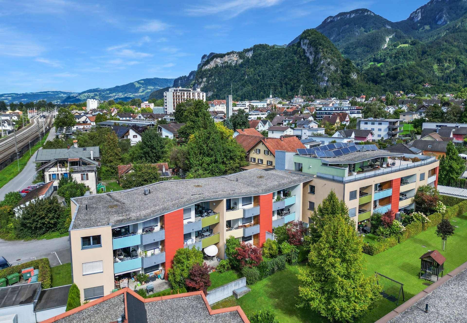 Mehrfamilienhaus mit Balkonen, Grünflächen und Bergblick unter blauem Himmel.