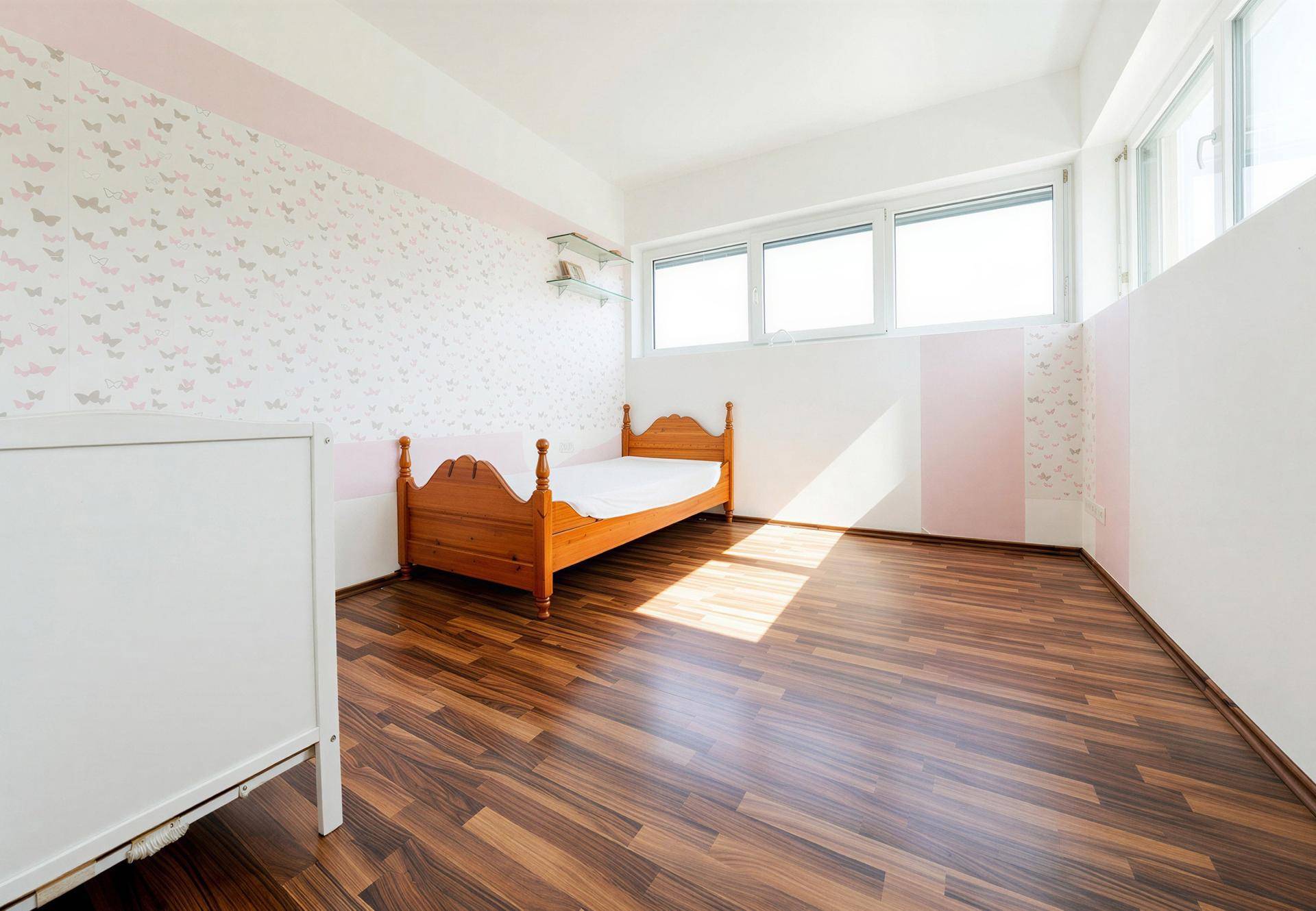 Kinderzimmer mit hellem Laminatboden, Fensterfront und verspielter rosa Schmetterlingstapete.
