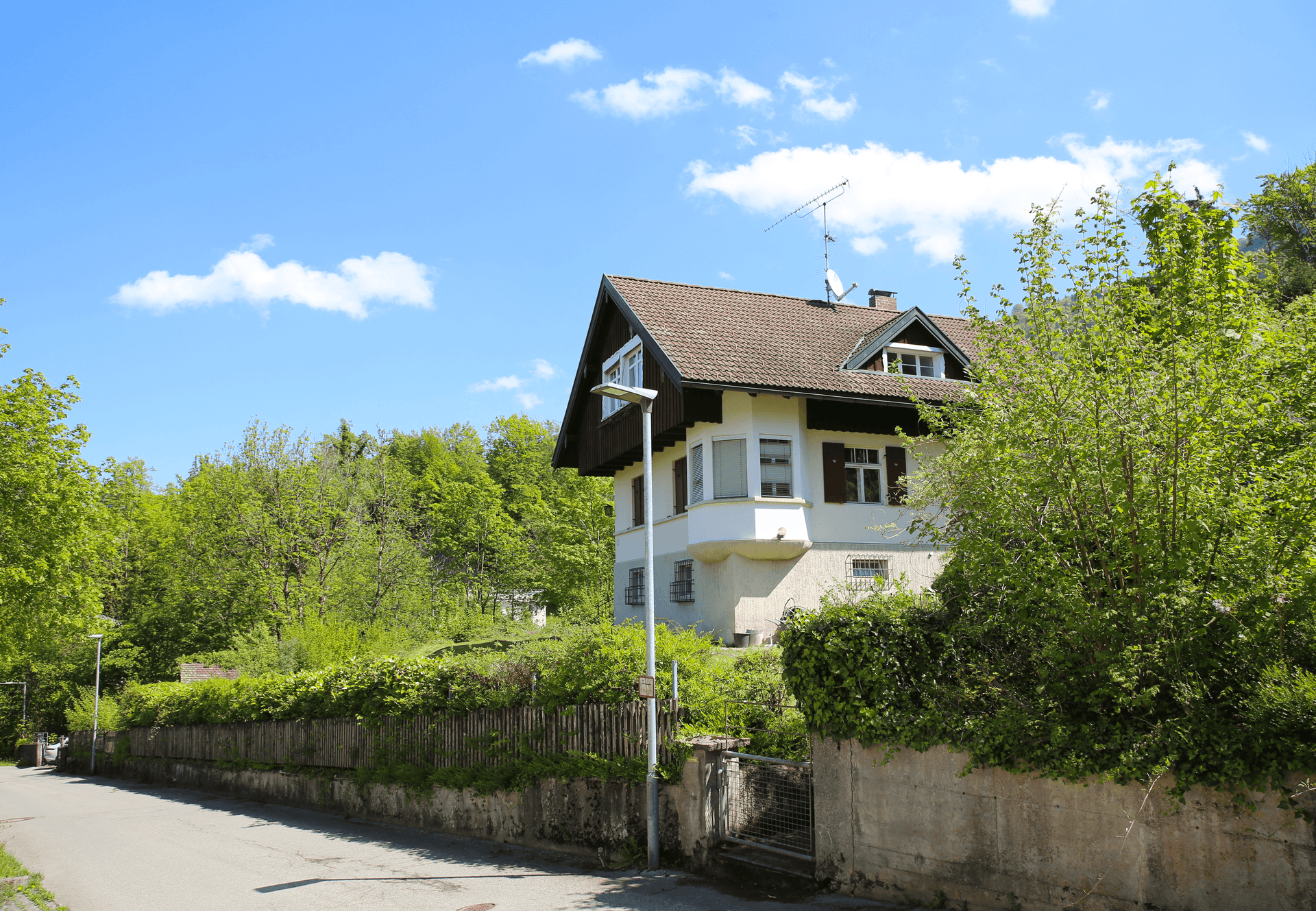 Haus von der Straße aus gesehen, umgeben von Bäumen und einem Holzzaun an einem sonnigen Tag.