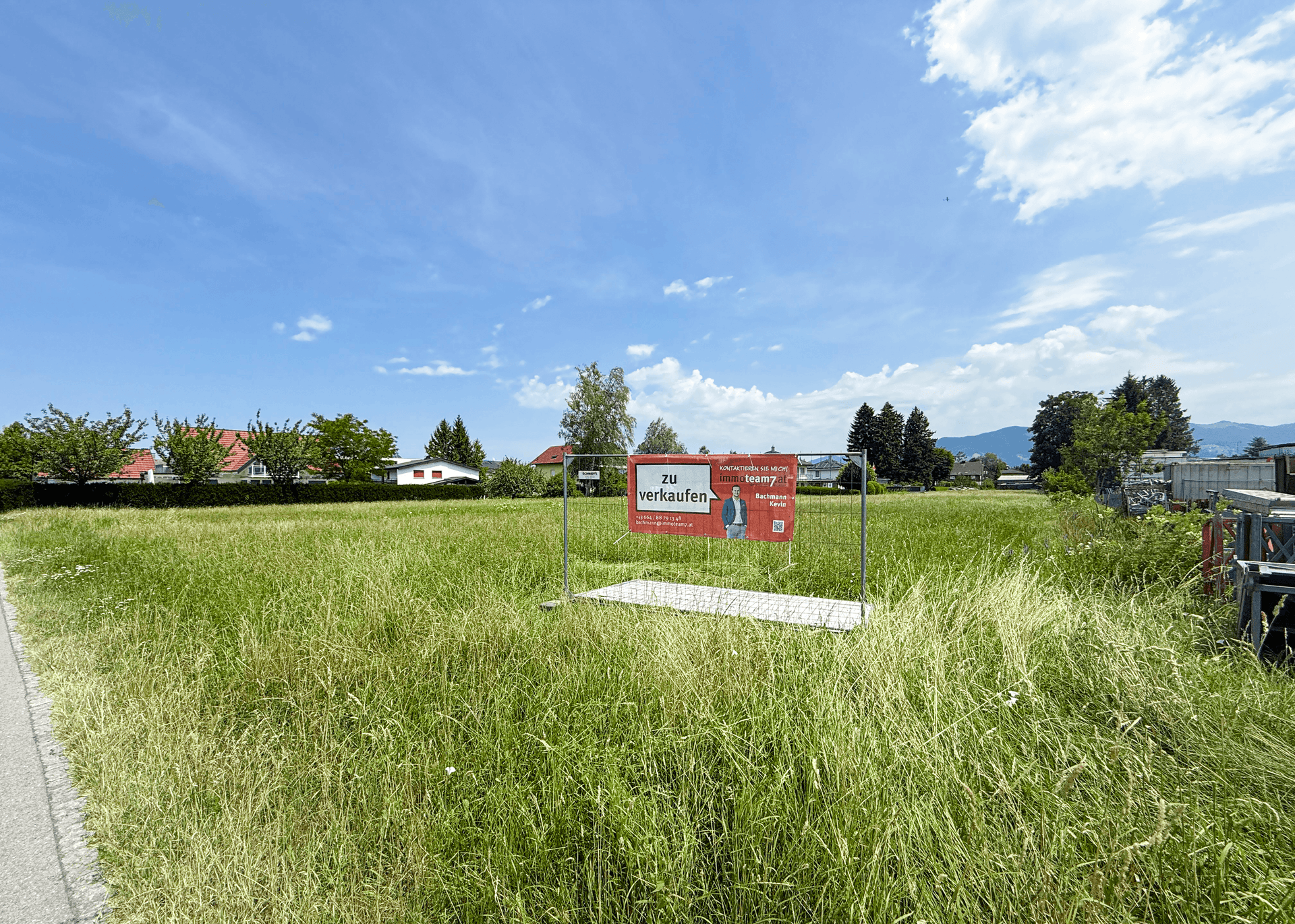 Blick vom Boden auf das grüne Baugrundstück mit 'Zu verkaufen'-Schild unter blauem Himmel.