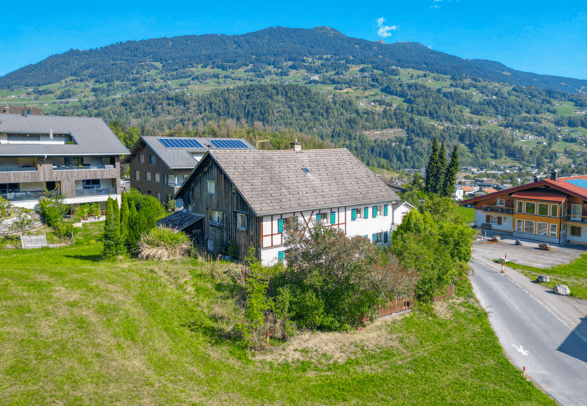Traditionelles Haus mit Holzelementen und grünen Wiesen, umgeben von Bergen und einer ländlichen Straße.