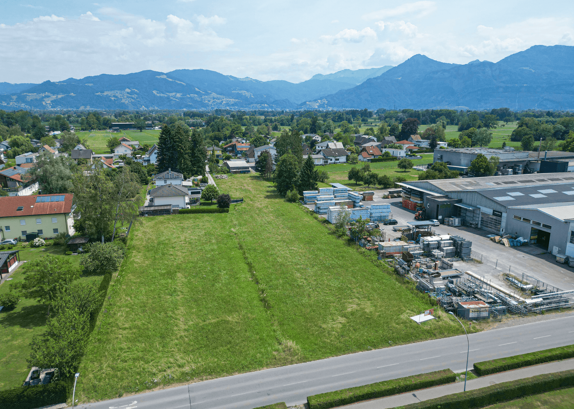 Luftaufnahme eines grünen Grundstücks neben einer Straße, umgeben von Wohn- und Industriegebäuden mit Bergblick.