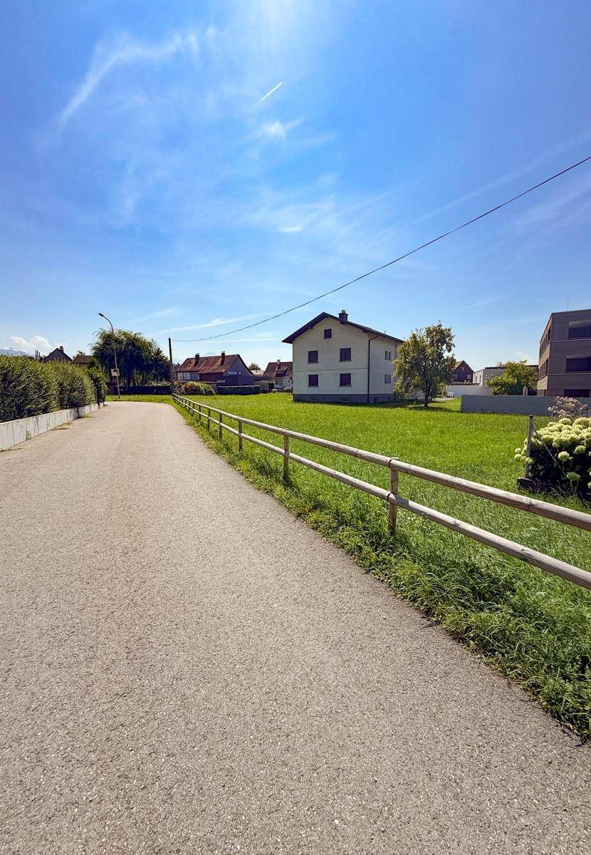 Asphaltierter Weg neben einer grünen Wiese mit einem traditionellen Haus und einem modernen Gebäude im Hintergrund.