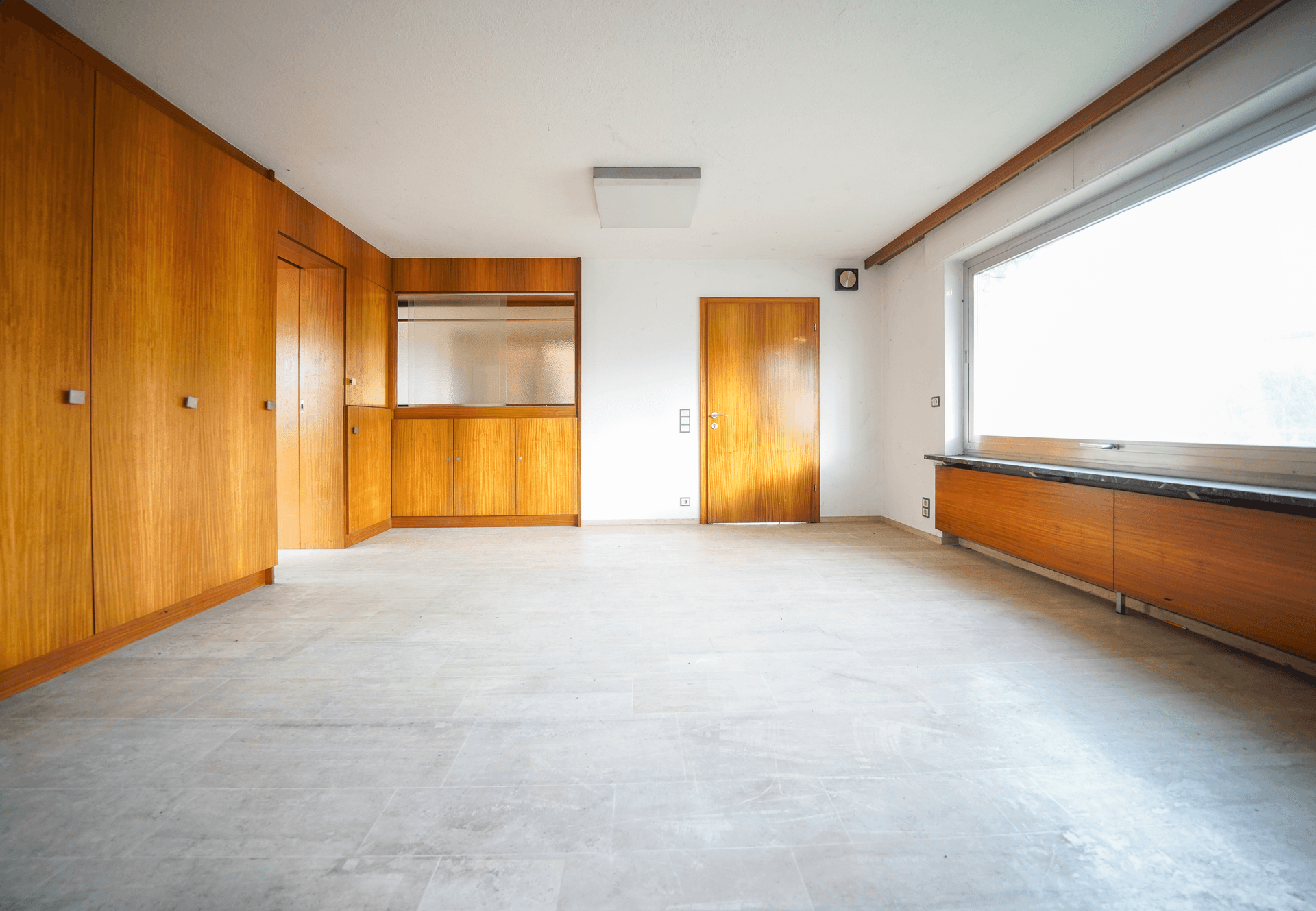 Großes, helles Zimmer mit Fliesenboden, Einbauschränken aus Holz und einem breiten Fenster.