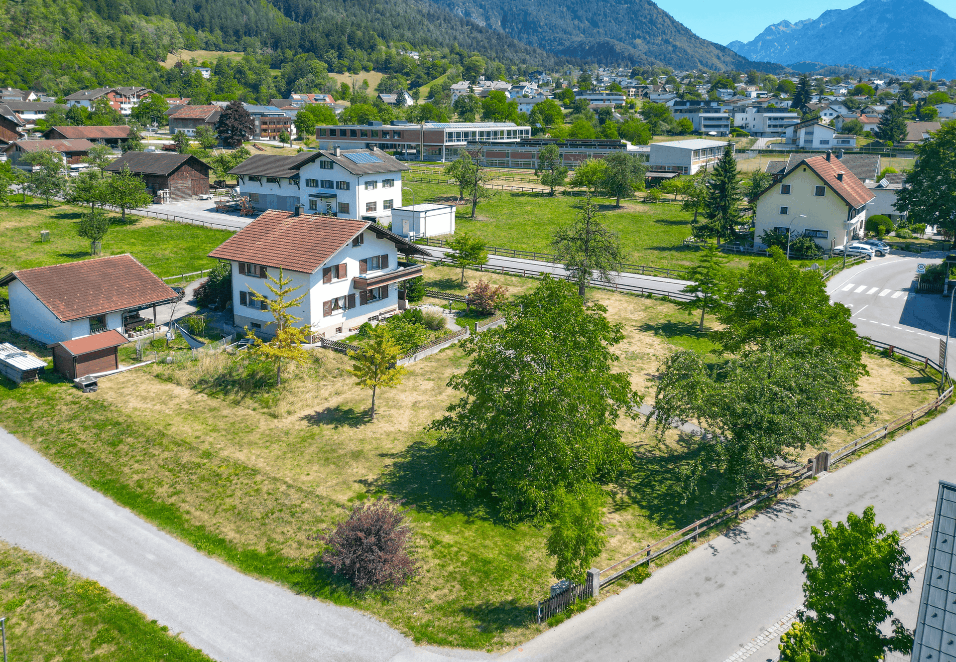 Blick von oben auf das Einfamilienhaus mit großem Garten und der umliegenden Siedlung vor alpiner Kulisse.