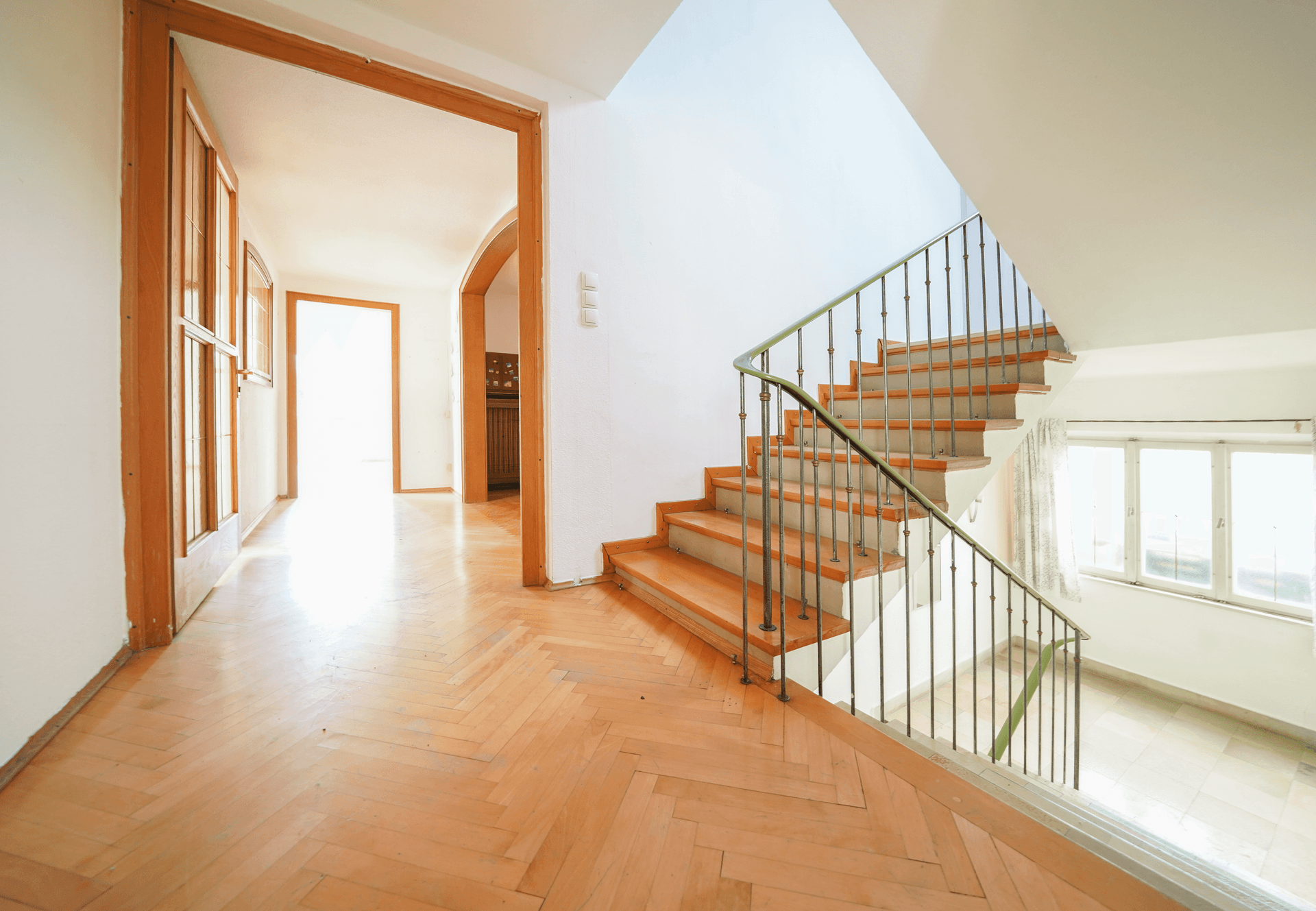 Heller Flur mit Fischgrätparkettboden und einer Treppe mit schmiedeeisernem Geländer.