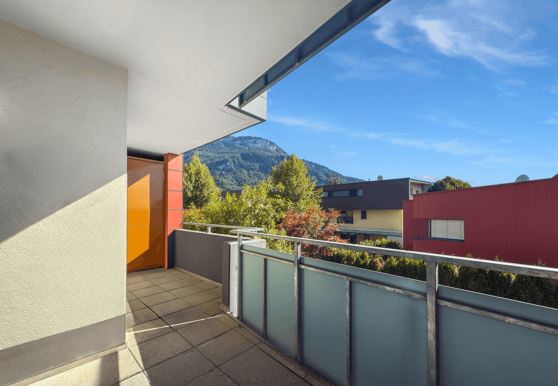 Balkon mit Bergblick, Fliesenboden und moderner Glasbrüstung unter blauem Himmel.