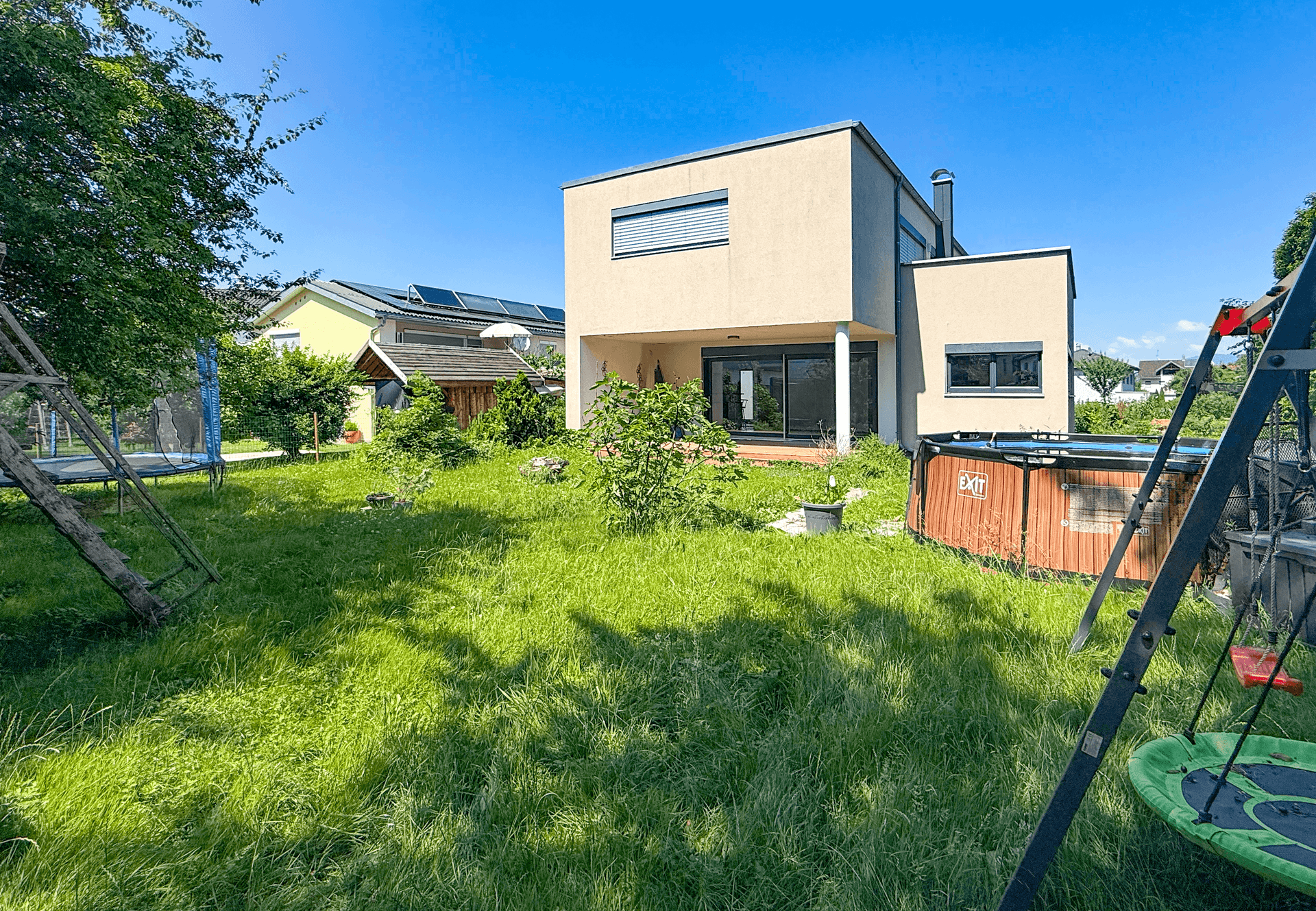 Großer, sonniger Garten mit Spielgeräten und einem modernen Haus im Hintergrund.