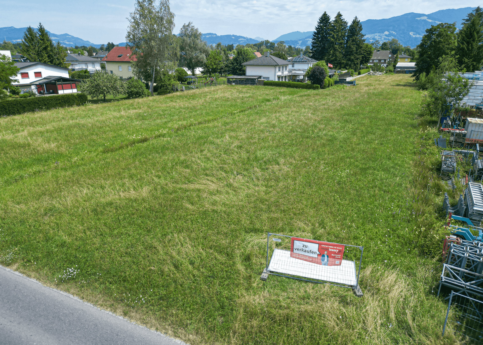 Grünes Grundstück mit einem 'Zu verkaufen'-Schild, umgeben von Wohnhäusern und Bergen im Hintergrund.