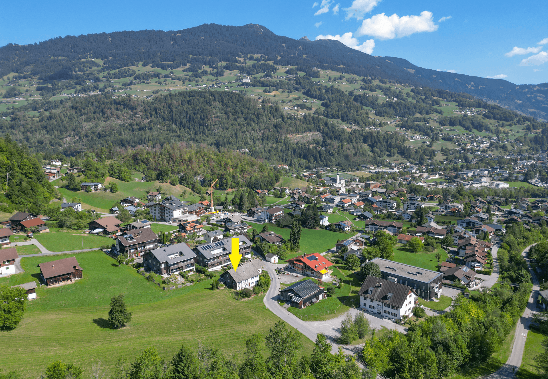 Panoramablick aus der Luft auf das Dorf und die umliegende Bergwelt, das Objekt ist mit einem Pfeil markiert.