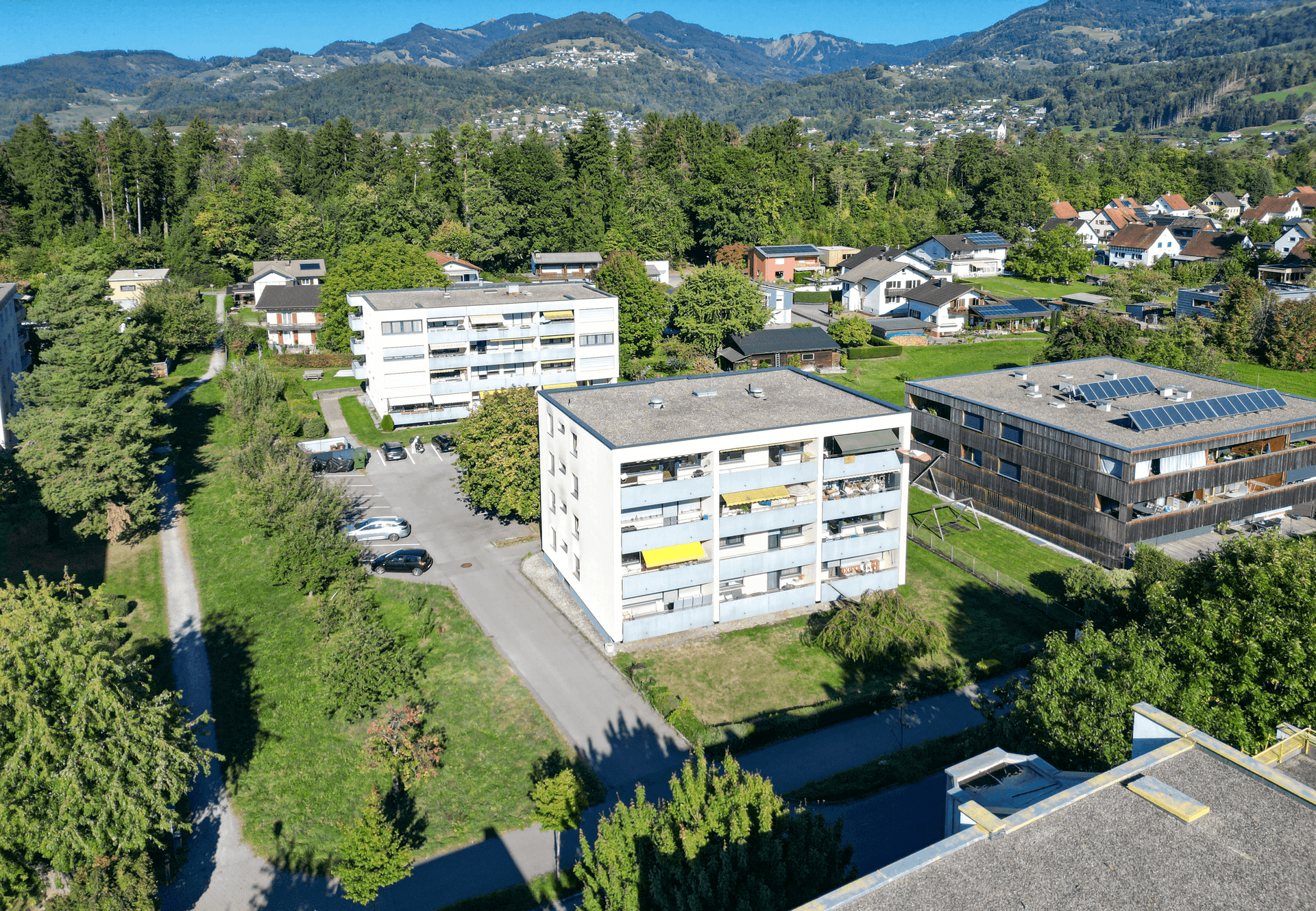 Wohngebäude in einer Siedlung mit viel Grün und Bergblick unter blauem Himmel.