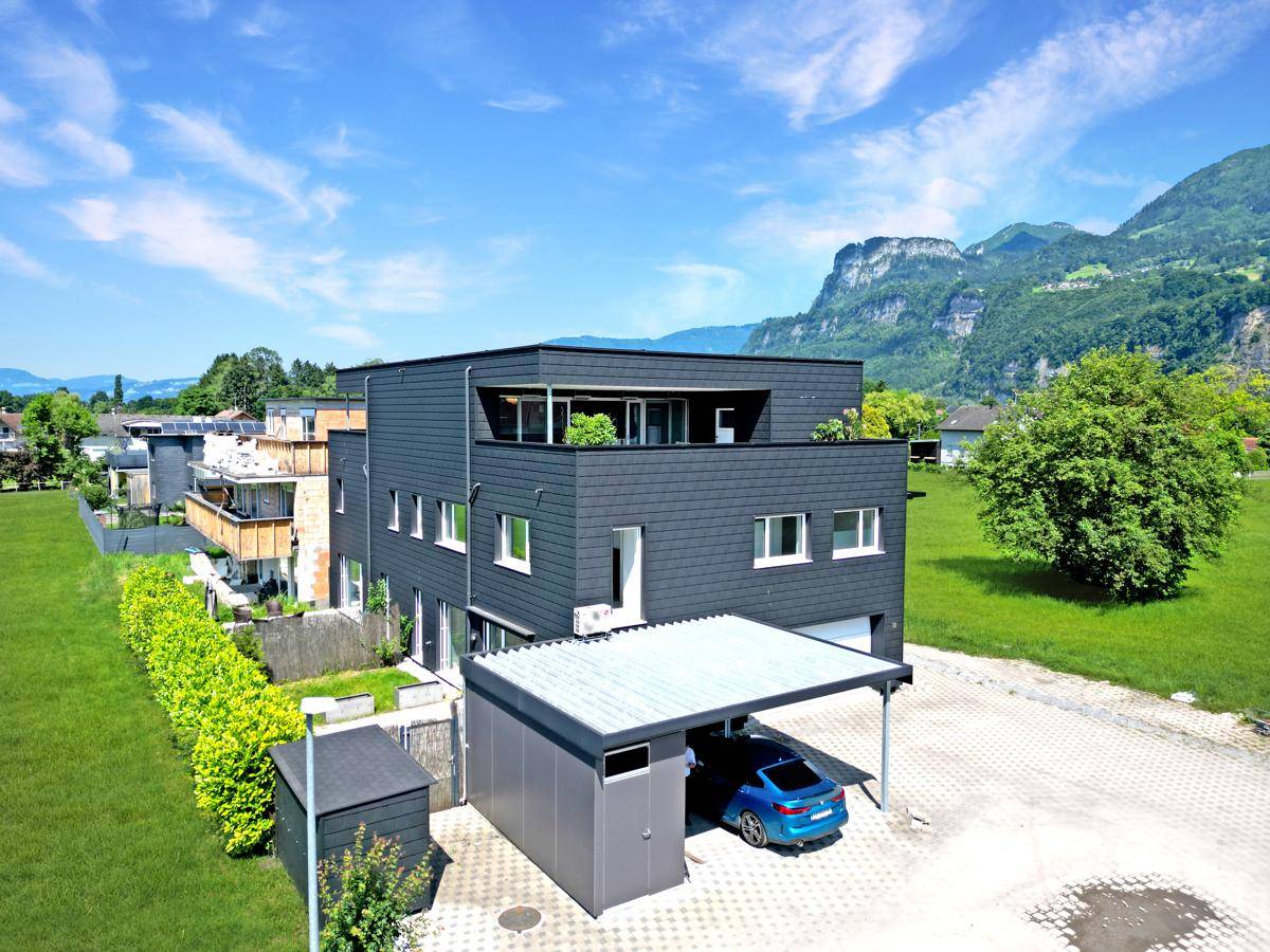 Modernes Mehrfamilienhaus mit dunkler Fassade, umgeben von grünen Wiesen und Bergen im Hintergrund.