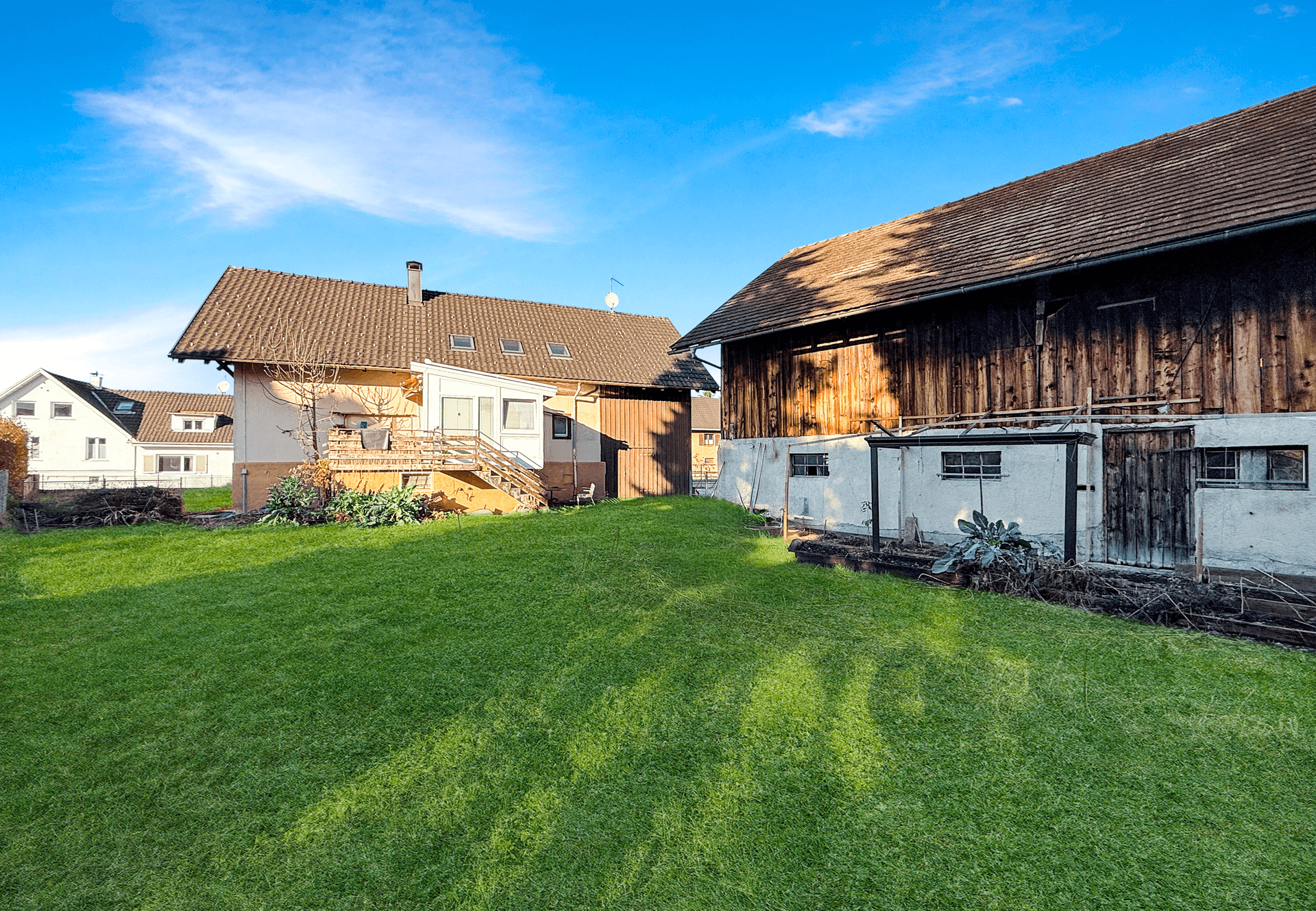 Großer grüner Gartenbereich mit einem Wohnhaus und einem rustikalen Holzgebäude unter blauem Himmel.