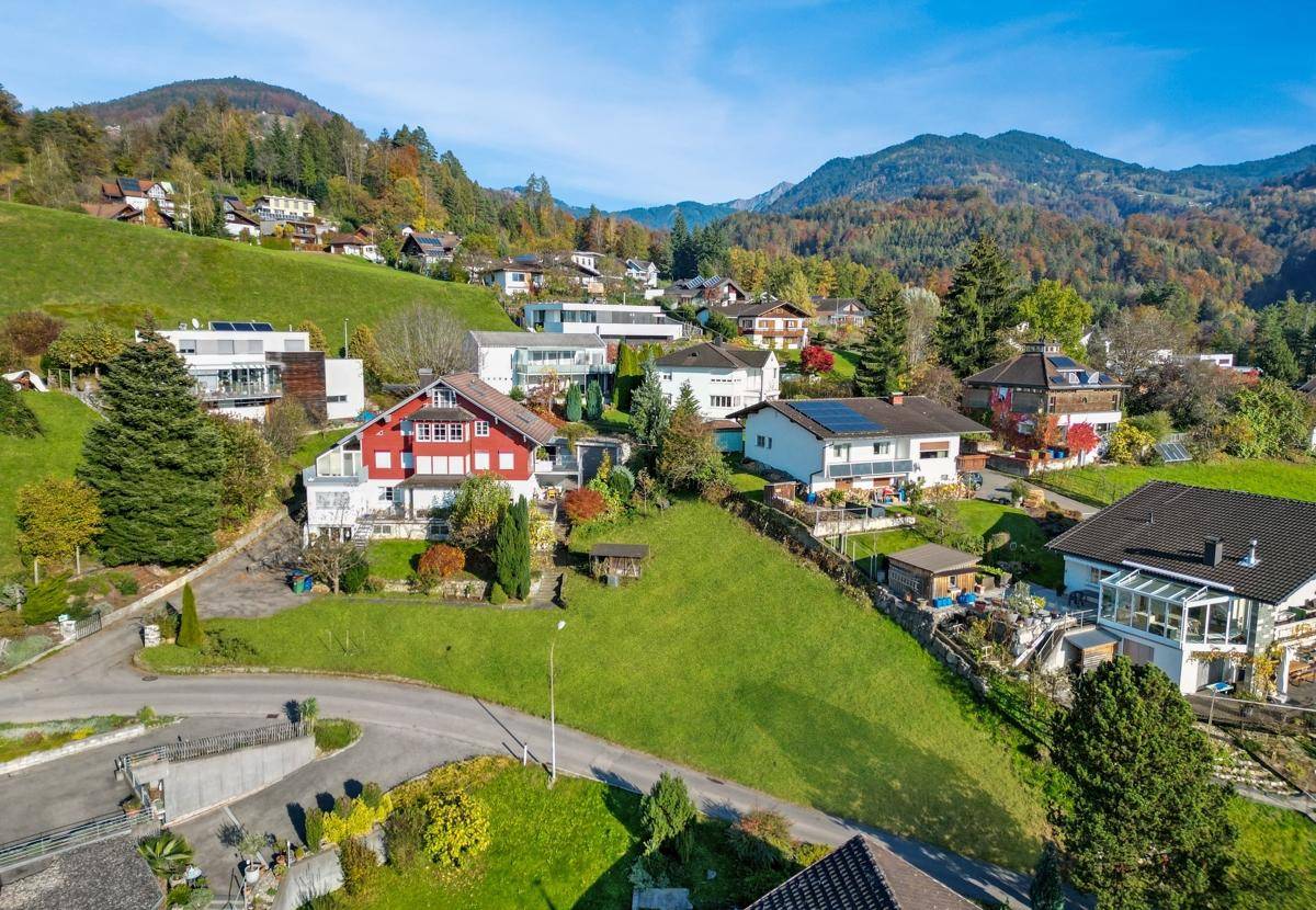 Luftaufnahme einer Wohnsiedlung in einem Tal, umgeben von bewaldeten Hügeln und Bergen.