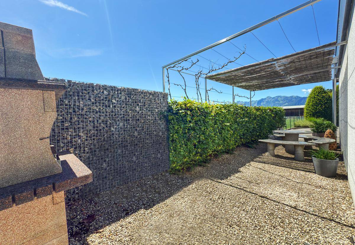 Gartenbereich mit Steingrill, Gabionenwand, Hecke und einem überdachten Sitzplatz unter einer Pergola.