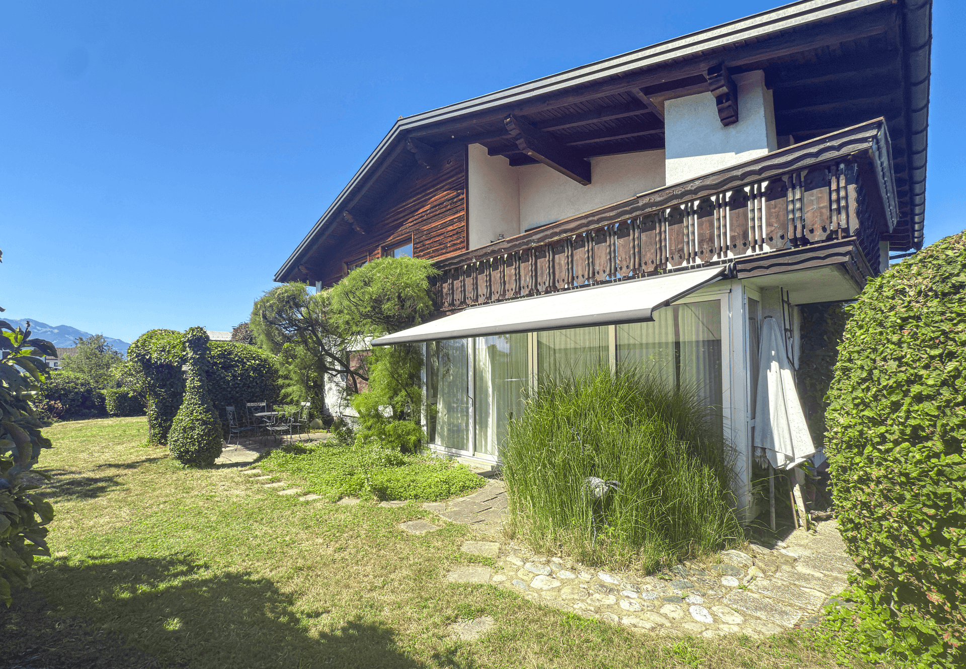 Sonniger Garten mit Rasenfläche, Wintergarten und traditionellem Balkon am Haus.