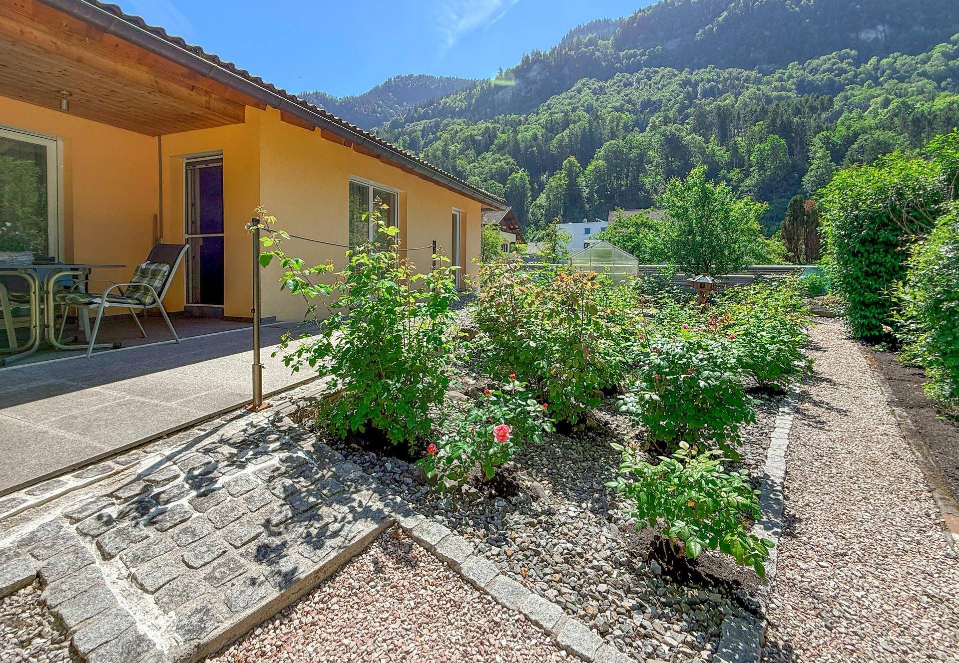 Gepflegter Garten mit Terrasse, Rosenbeeten und Kieswegen vor Bergkulisse.