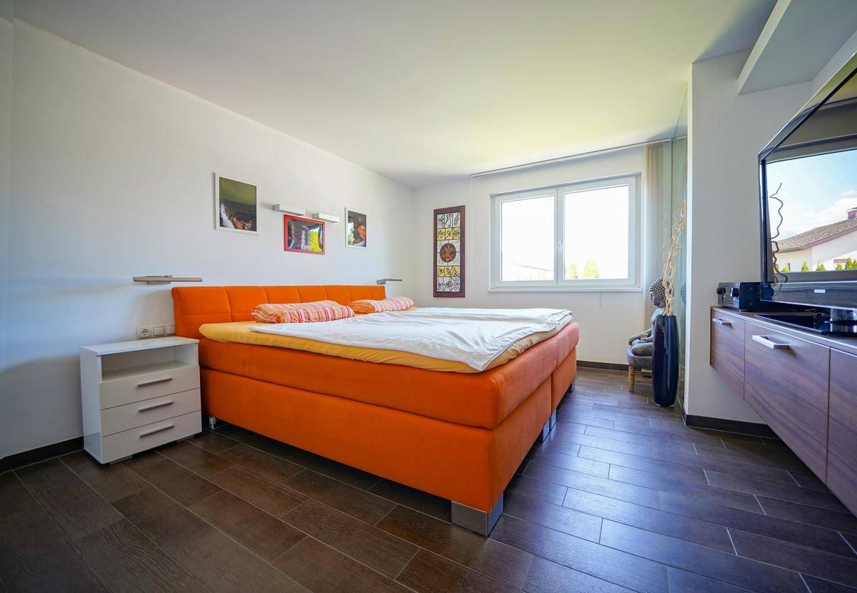 Geräumiges Schlafzimmer mit orangefarbenem Boxspringbett, Fenster und dunklem Fliesenboden.