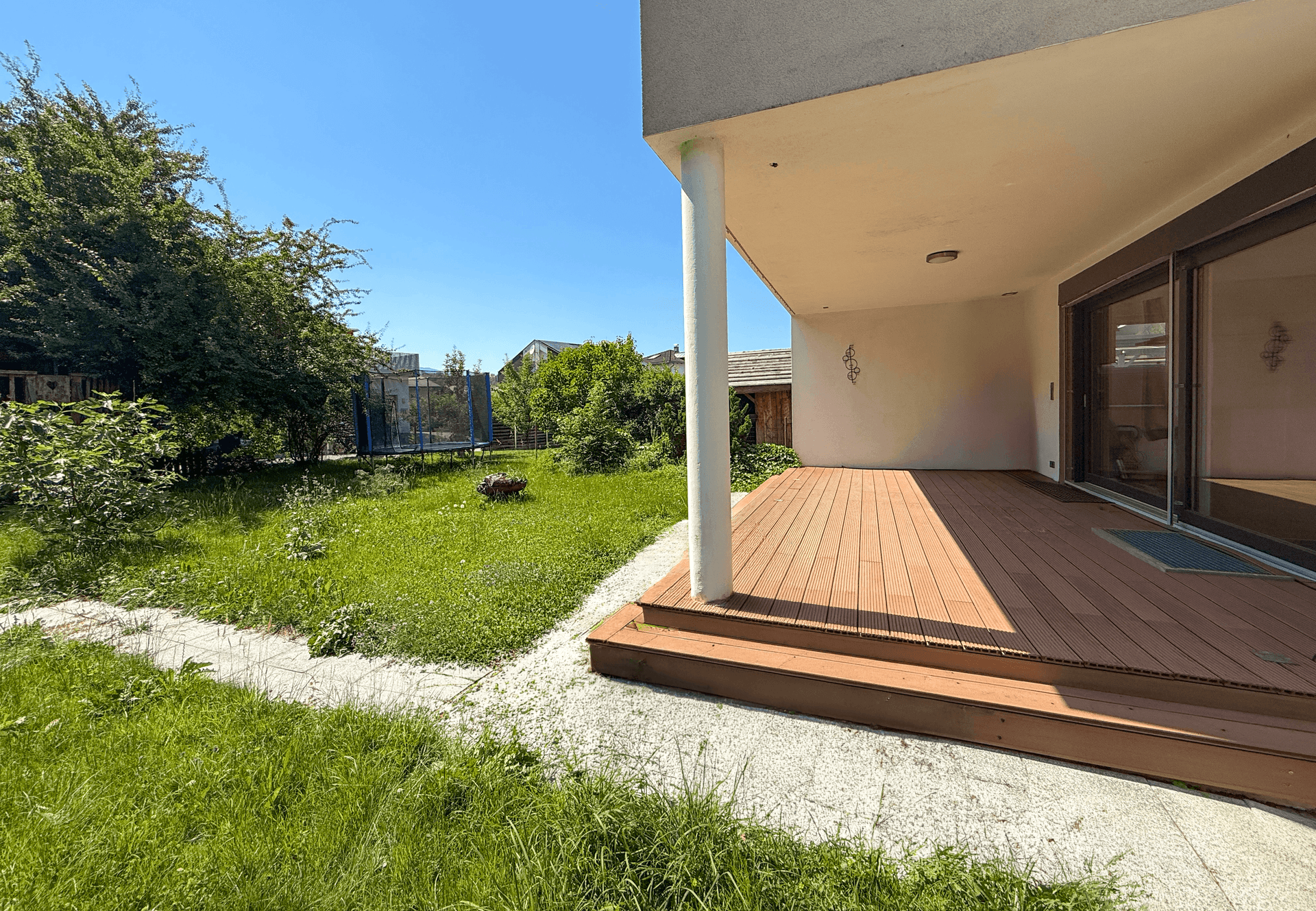 Sonniger Terrassenbereich mit Holzboden und Zugang zum weitläufigen grünen Garten.