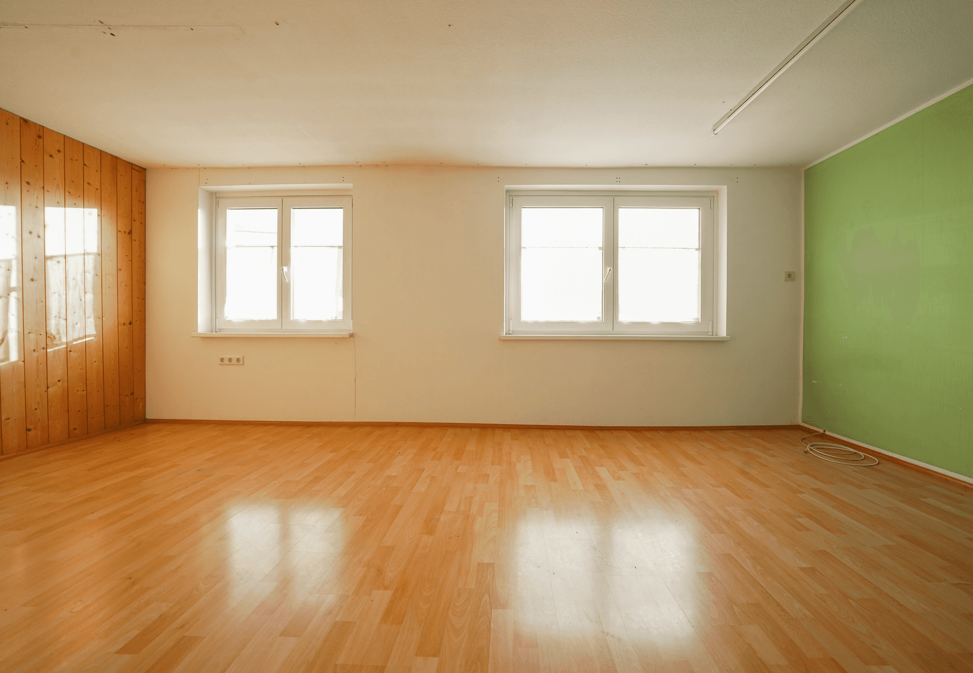 Geräumiges Zimmer mit Holzwandverkleidung, grüner Akzentwand und zwei großen Fenstern.