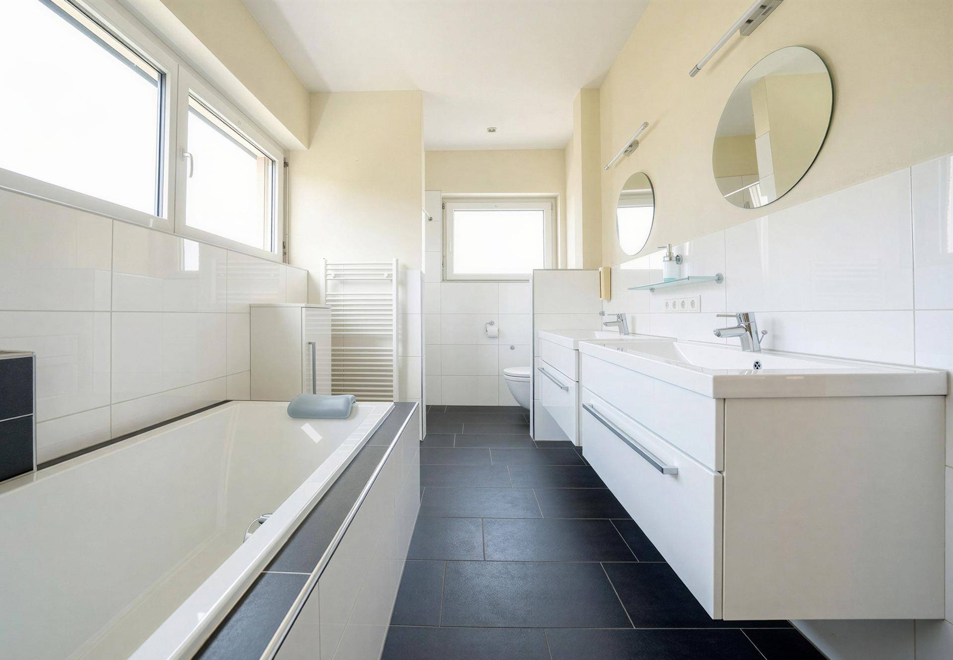 Modernes Badezimmer mit Doppelwaschbecken, Badewanne, Fenster und dunklen Fliesen.