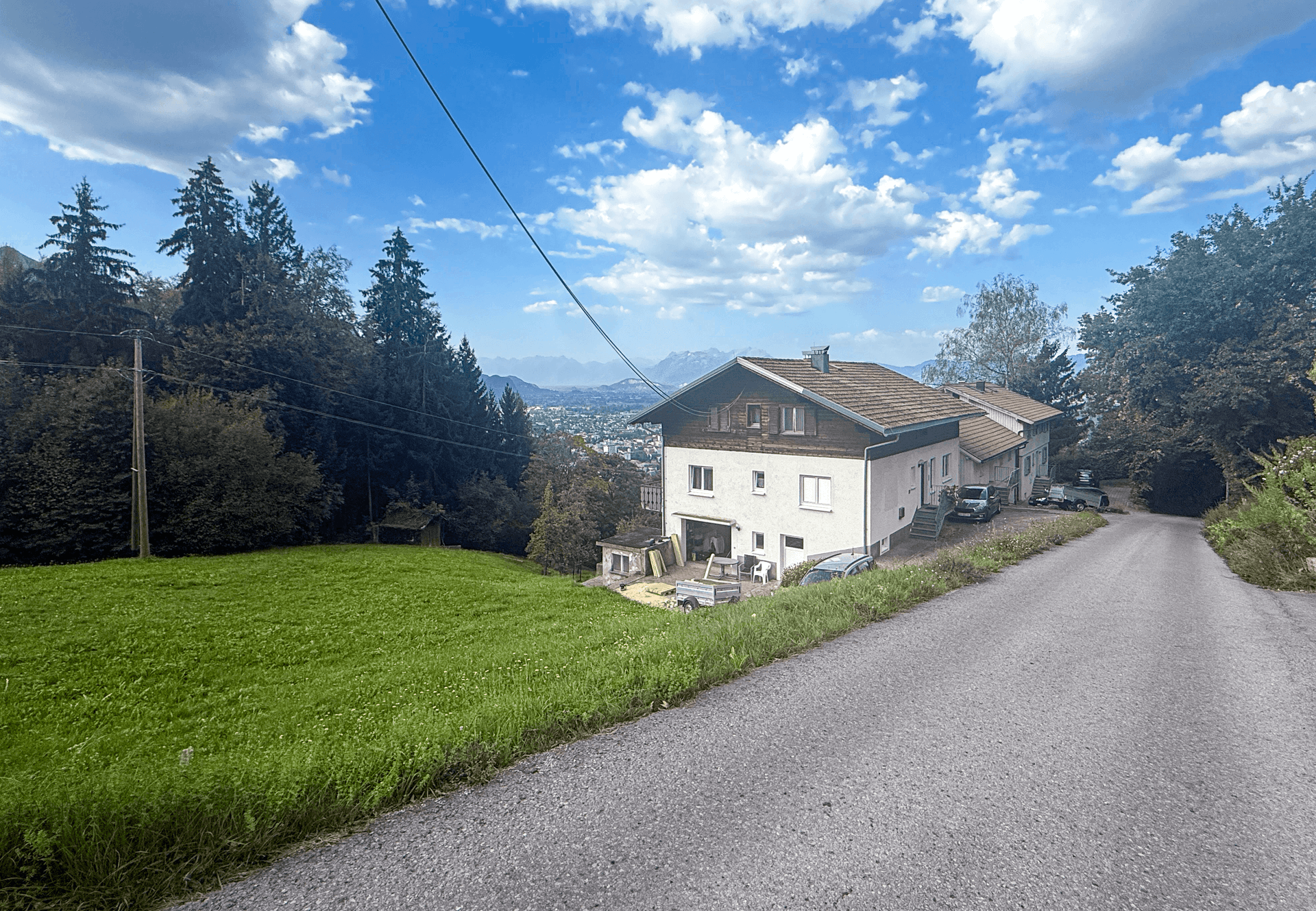Landstraße führt an einem Wohnhaus vorbei, umgeben von einer grünen Wiese und mit Fernblick auf Stadt und Berge.