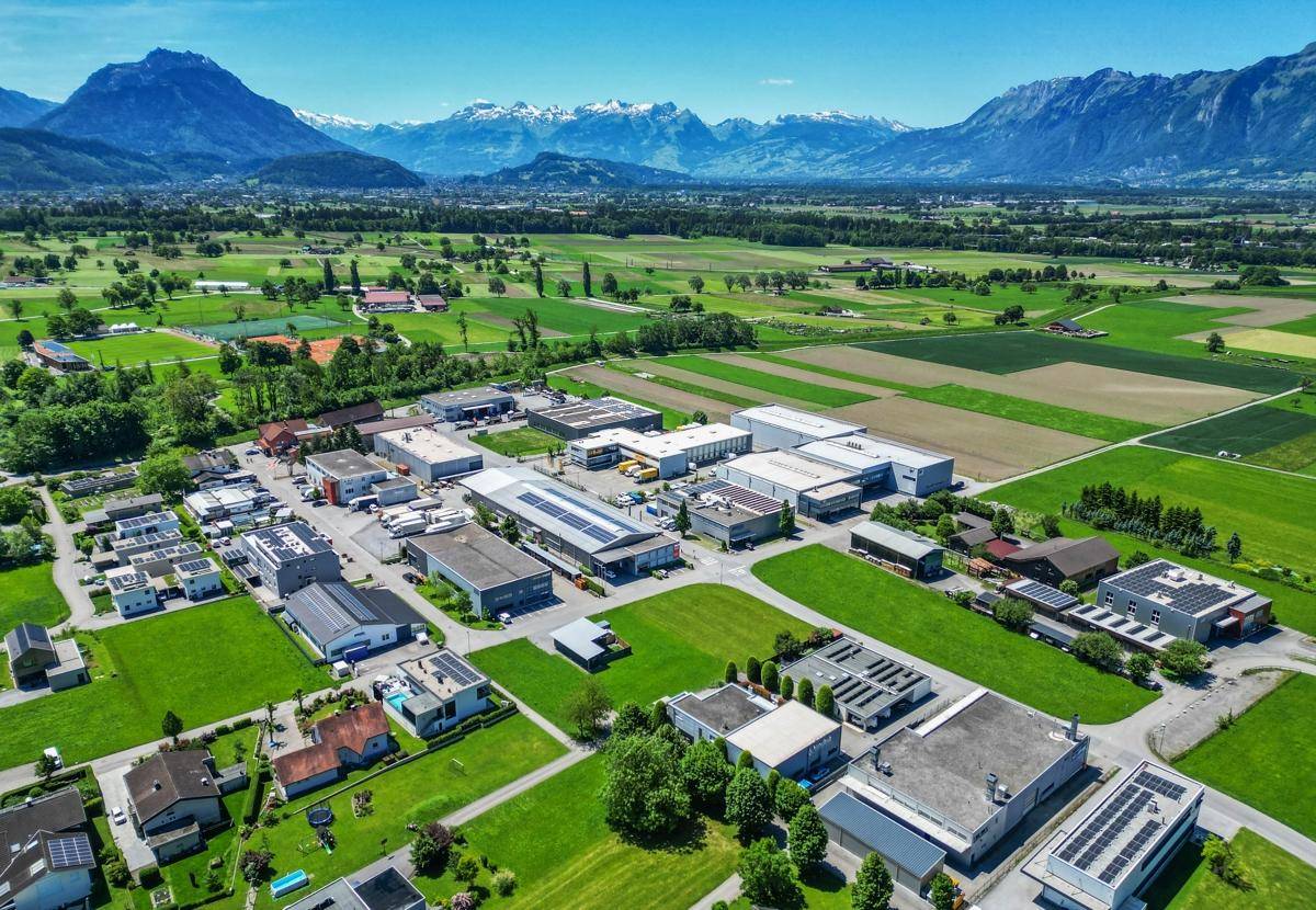 Luftaufnahme eines Gewerbegebiets mit vielen Gebäuden, grünen Feldern und schneebedeckten Bergen.