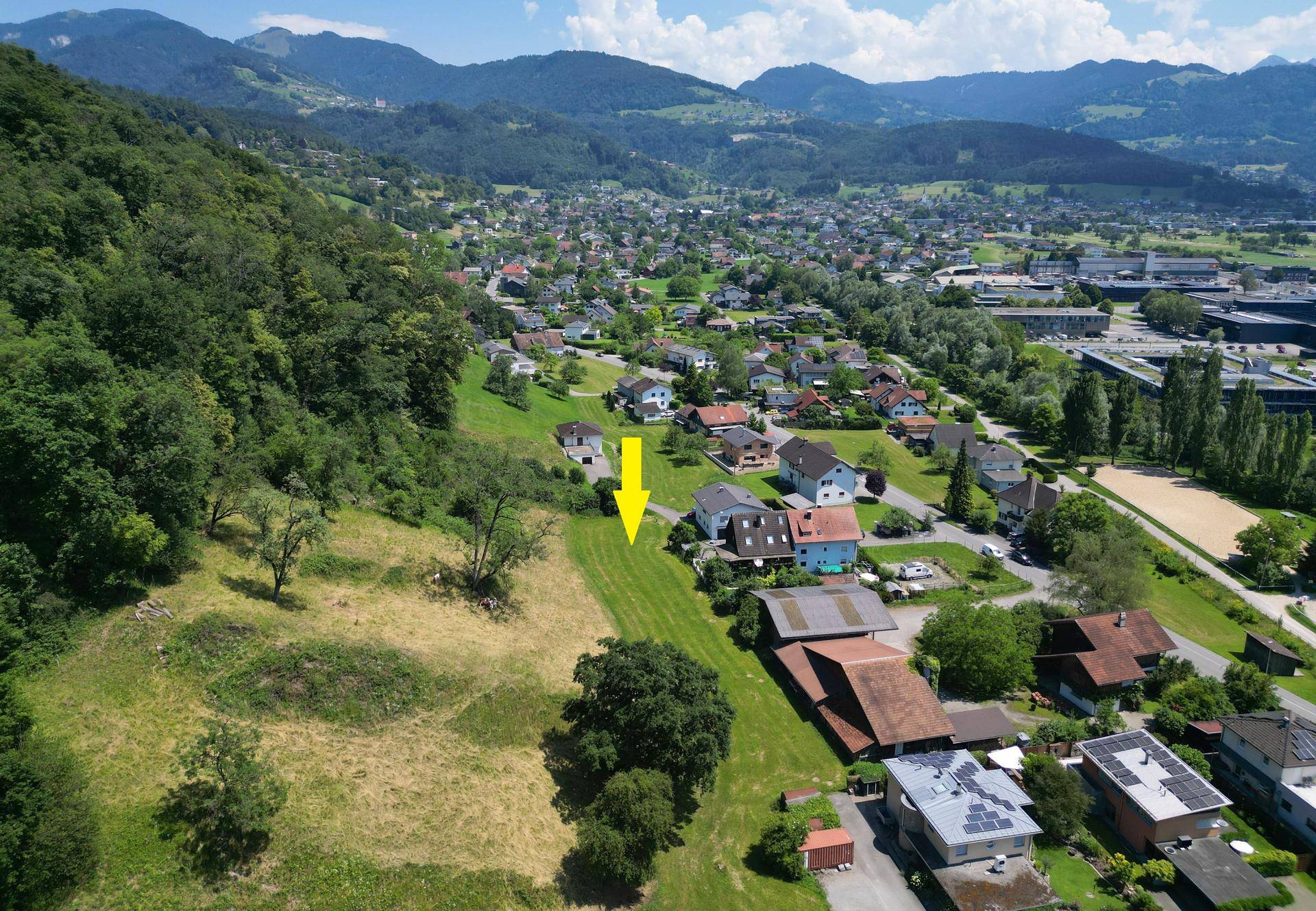 Luftaufnahme einer Siedlung mit Häusern, Feldern und einem Waldgebiet am Hang.