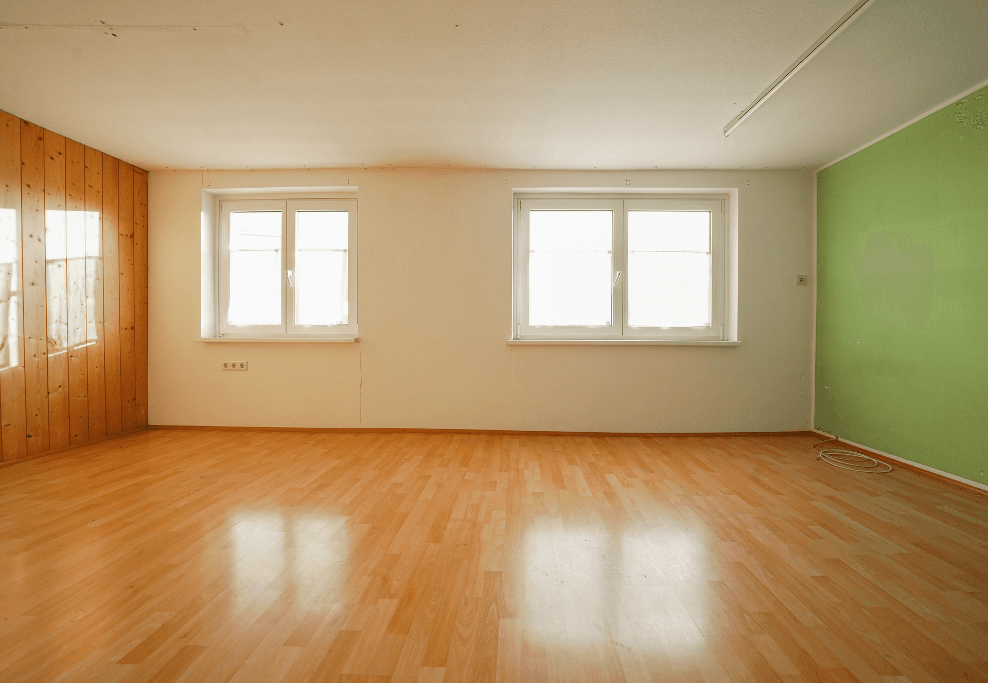 Leeres Zimmer mit hellem Holzfußboden, zwei Fenstern und einer grünen Akzentwand.