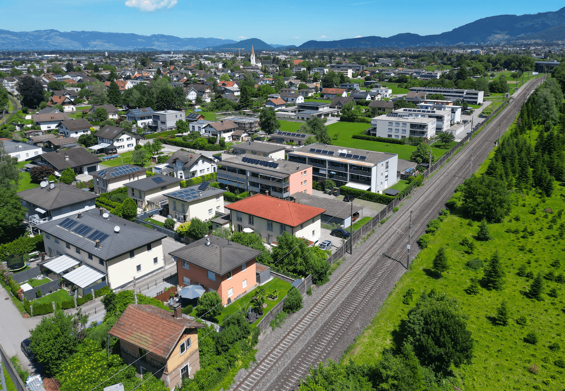 Weitläufige Luftaufnahme der Wohnsiedlung mit Bahngleisen und Bergpanorama im Hintergrund, sehr idyllisch.