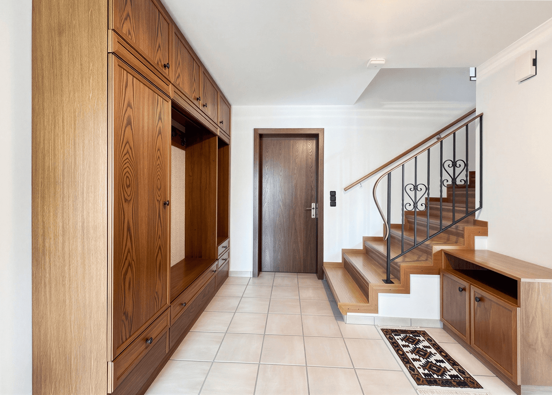 Eingangsbereich mit Fliesenboden, Holzschränken und Treppe mit schmiedeeisernem Geländer.