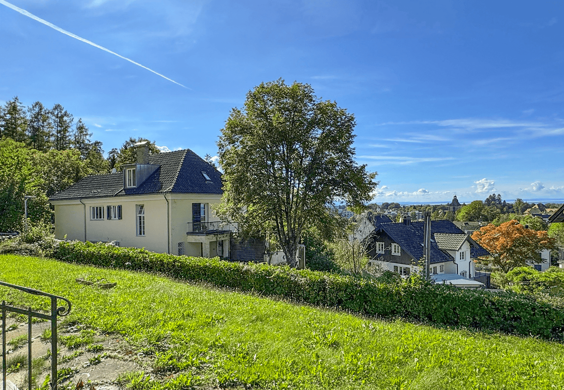 Blick von einem grünen Hang auf ein Nachbarhaus mit großem Baum und See im Hintergrund.