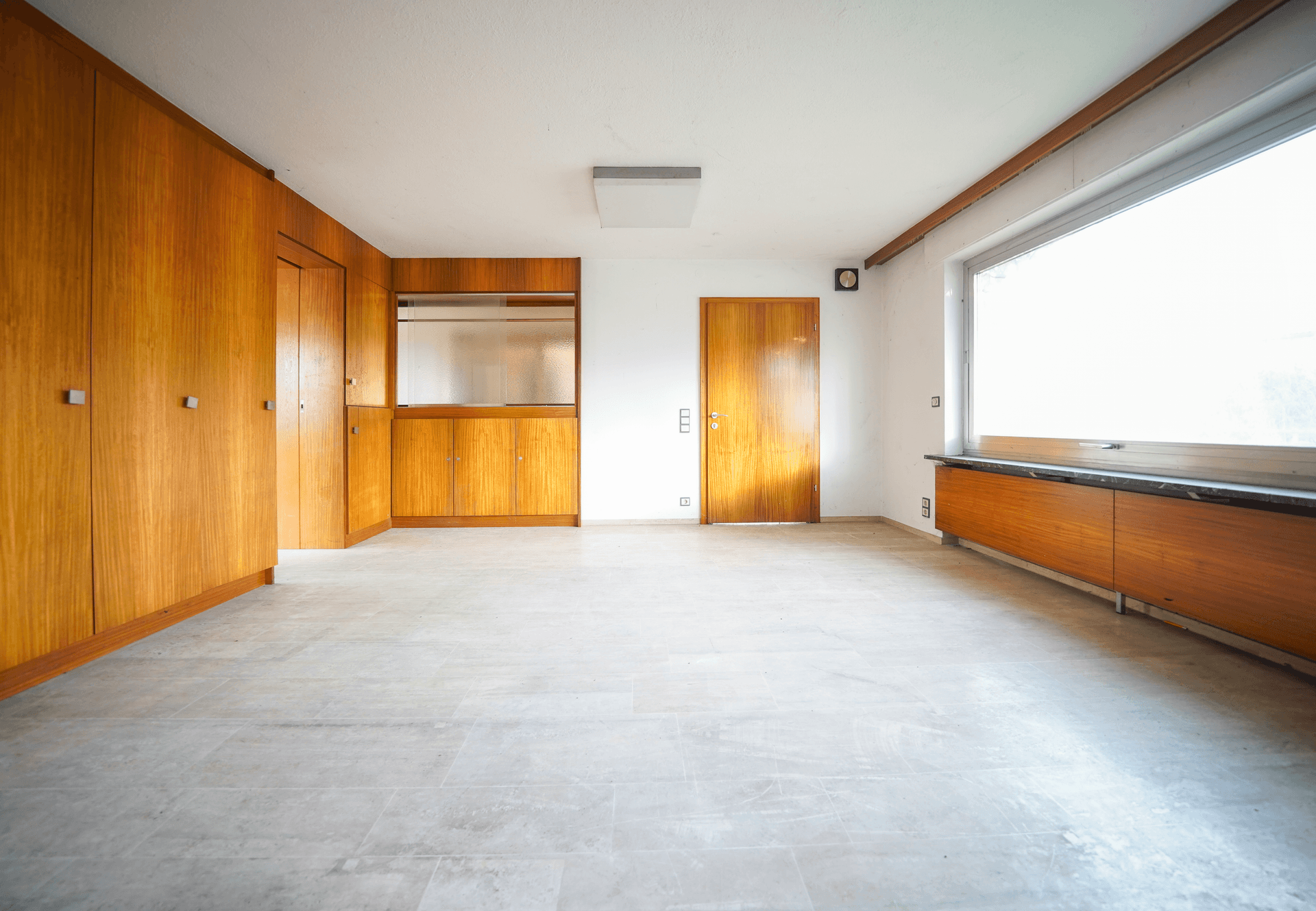 Geräumiges Zimmer mit hellen Fliesen, Einbauschränken aus Holz und einem großen Fenster.