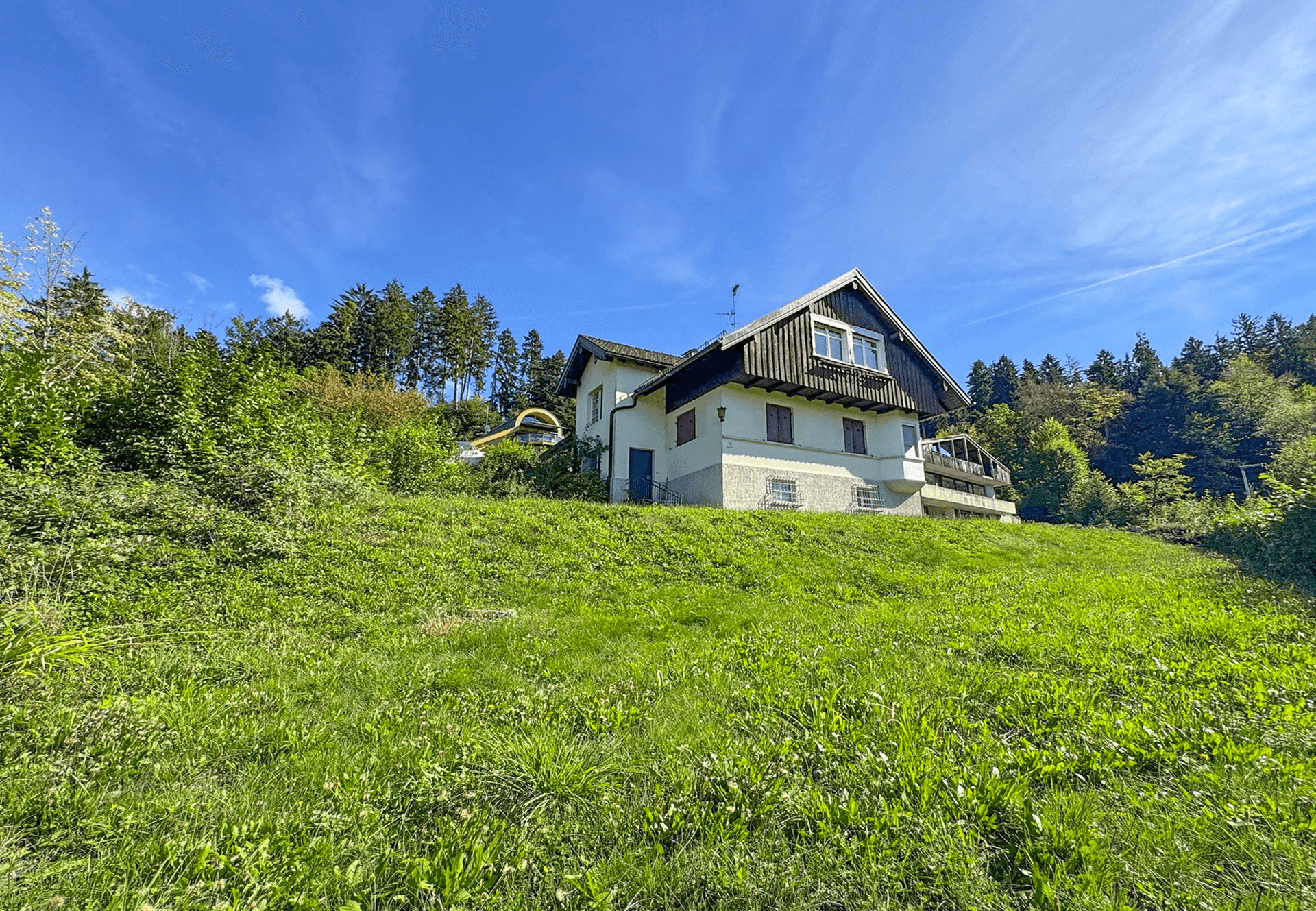 Haus auf einem grünen Hanggrundstück mit Wald im Hintergrund unter strahlend blauem Himmel.