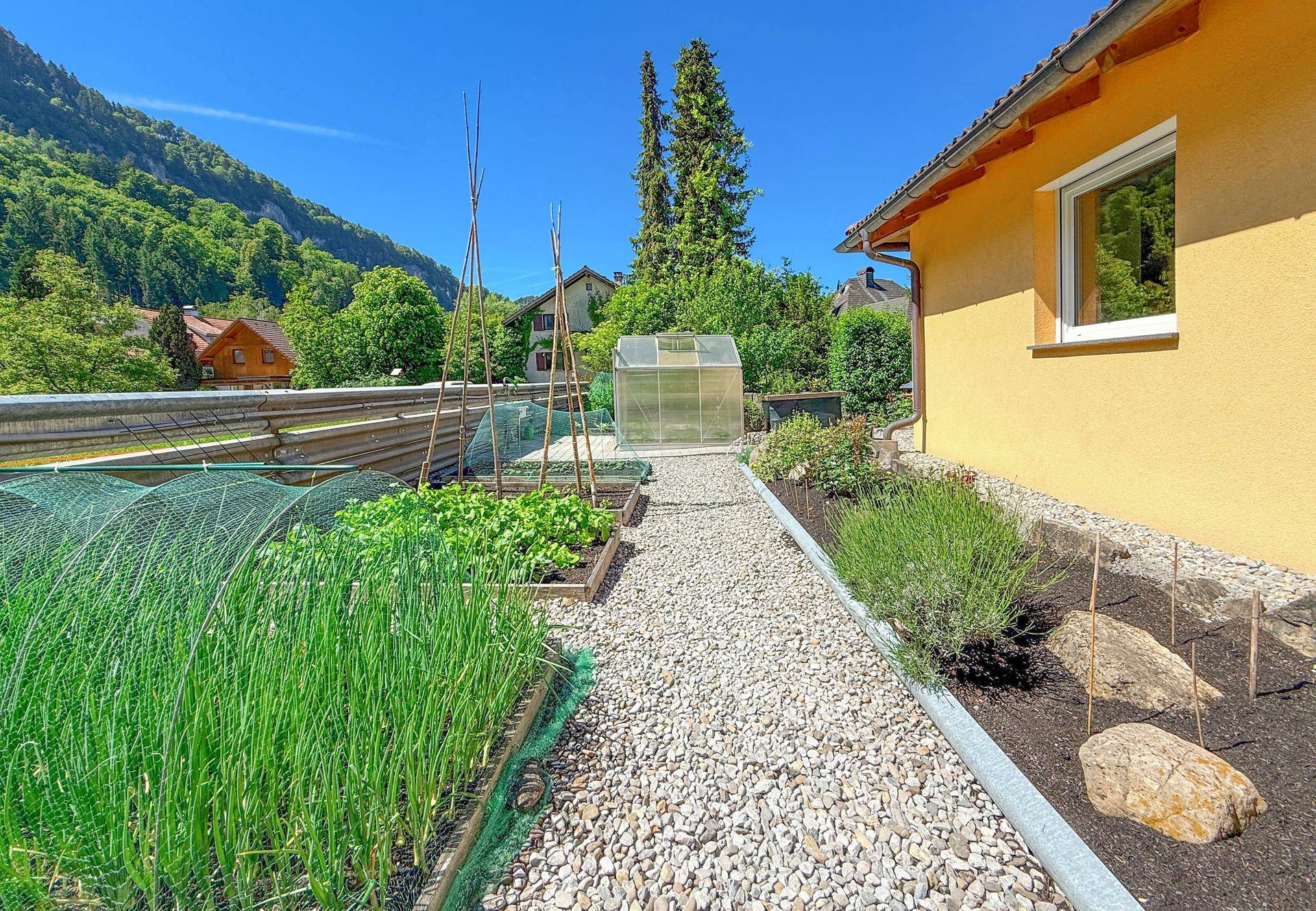 Funktionaler Gemüsegarten mit Hochbeeten, Gewächshaus und Kieswegen neben dem Haus.