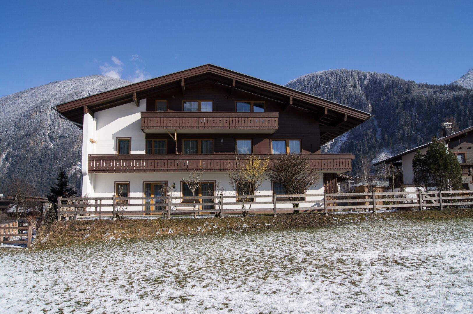 Traditionelles Haus im alpinen Stil mit Balkonen und schneebedeckter Berglandschaft im Hintergrund.