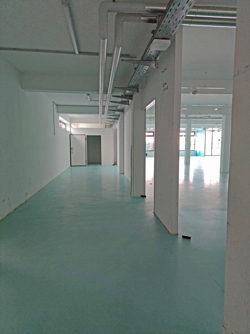 Langer Flur im Gewerbebereich mit hellblauem Boden, sichtbaren Leitungen und mehreren Türen.