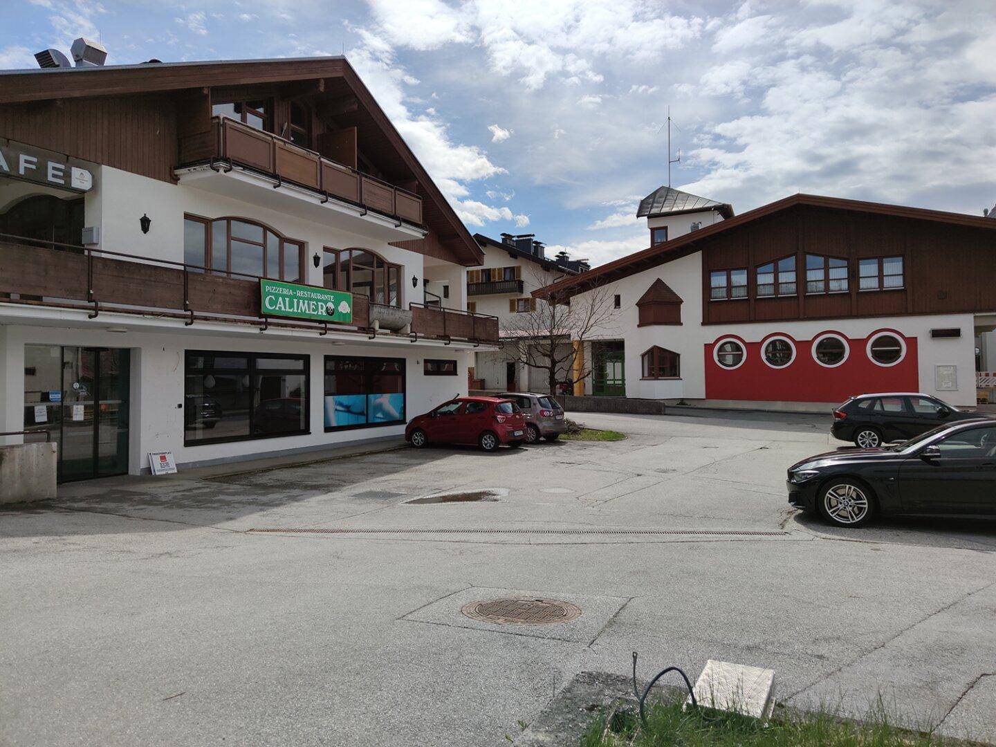 Gebäude mit Pizzeria und Wohnungen, umgeben von weiteren Häusern und Bergen unter bewölktem Himmel.