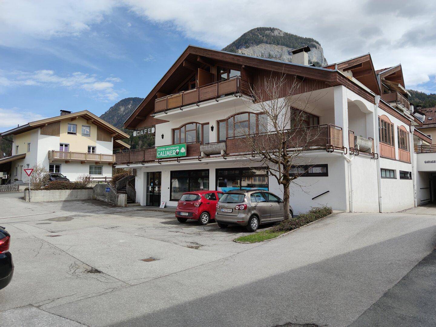 Vorderansicht des Gebäudes mit Pizzeria und Wohnungen, umgeben von Bergen und Parkplätzen.