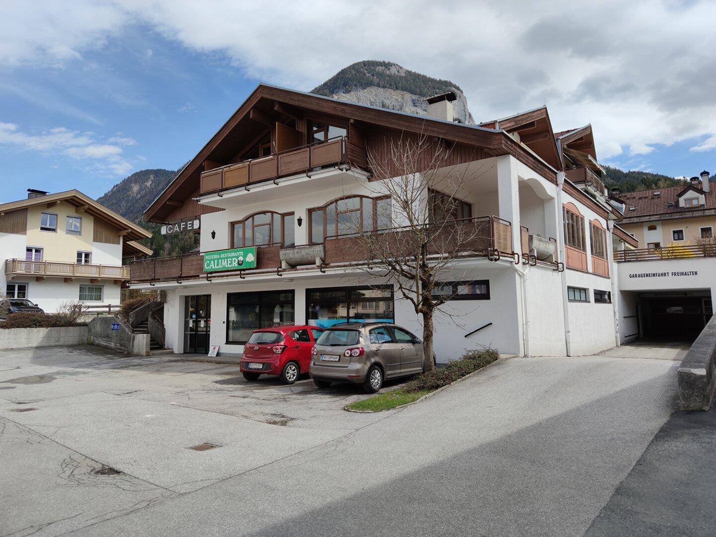 Gebäudeansicht mit Pizzeria, Wohnungen und einer Einfahrt zur Tiefgarage, im Hintergrund Berge.