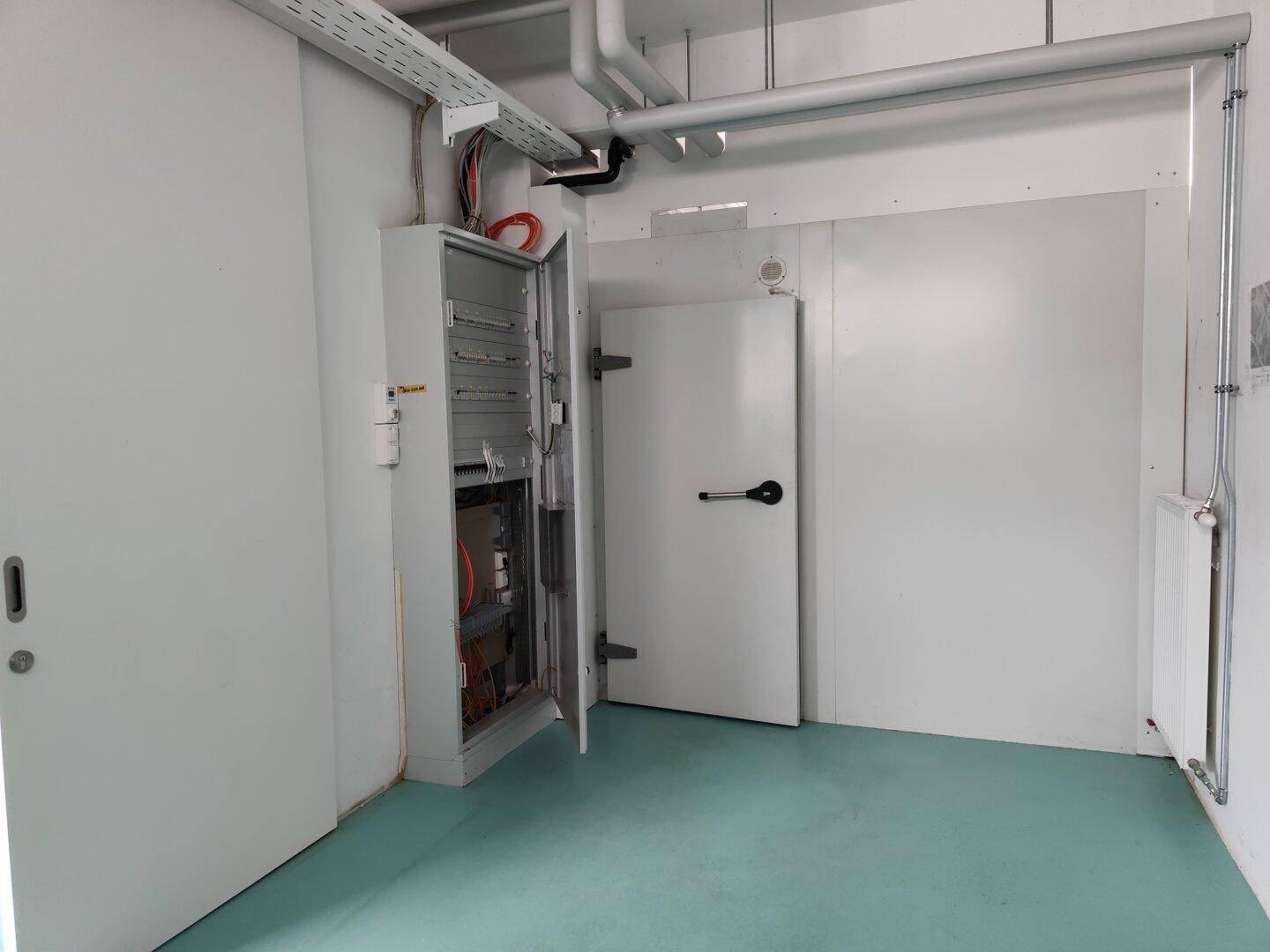 Technikraum mit offenem Elektroschrank, sichtbaren Leitungen und einer großen weißen Tür.