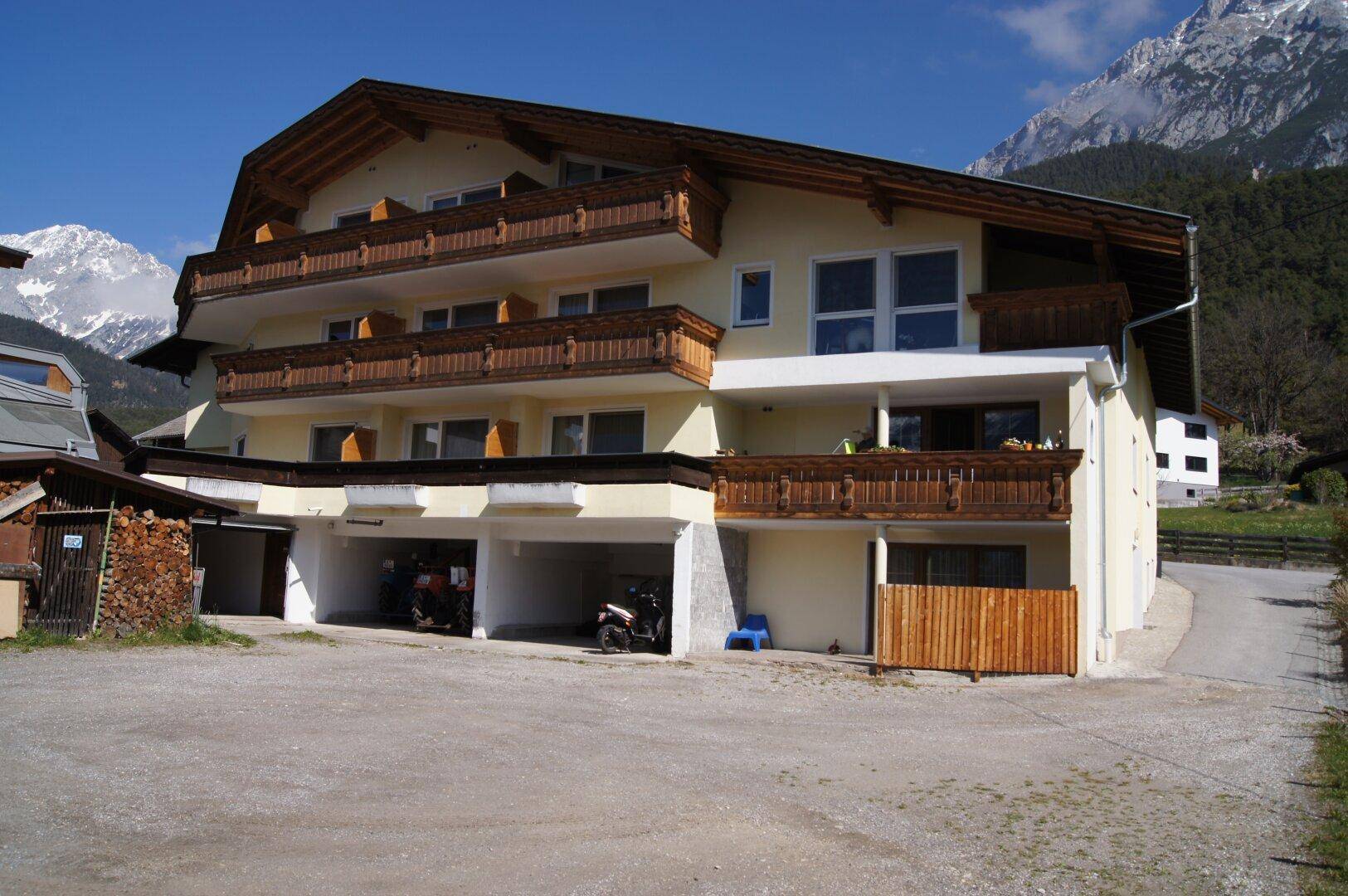 Traditionelles Mehrfamilienhaus mit Holzbalkonen und überdachten Parkplätzen vor Bergkulisse.