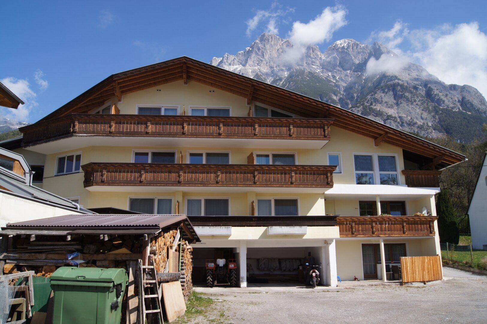 Mehrfamilienhaus mit gelber Fassade, Holzverkleidung und traditionellen Balkonen vor alpiner Kulisse.