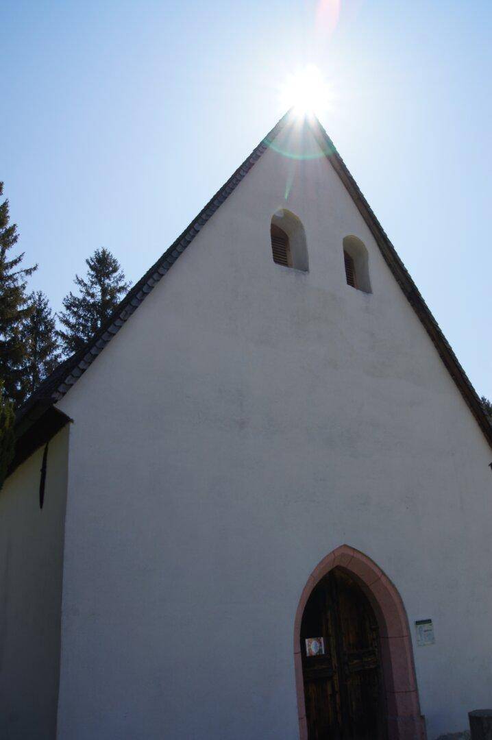 Historische Kapelle mit weißer Fassade und spitzem Dach, sonnenbeschienen vor blauem Himmel.