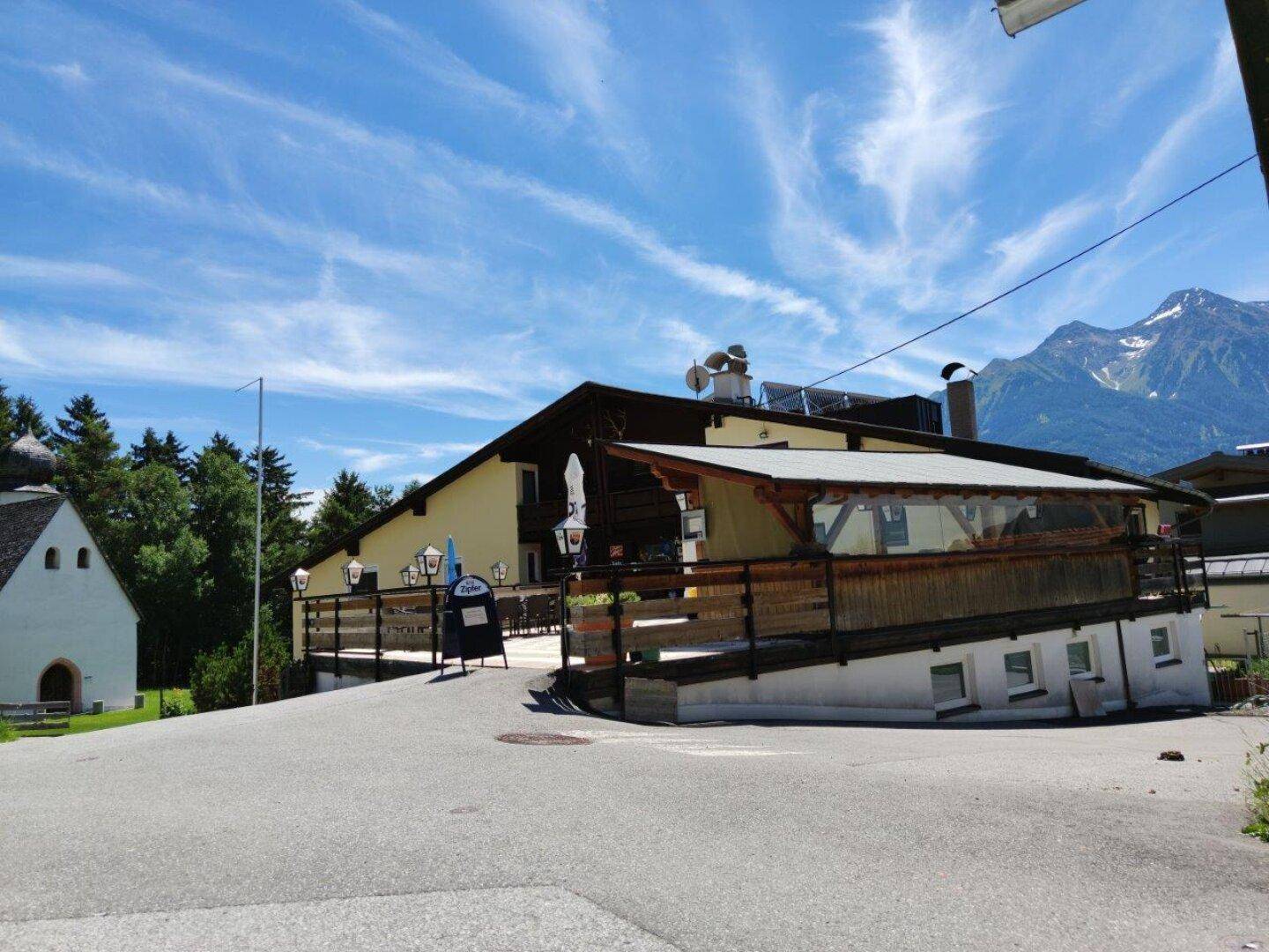 Gebäude mit Terrasse und Außenbereich, umgeben von alpiner Landschaft und einem kleinen Kirchlein.