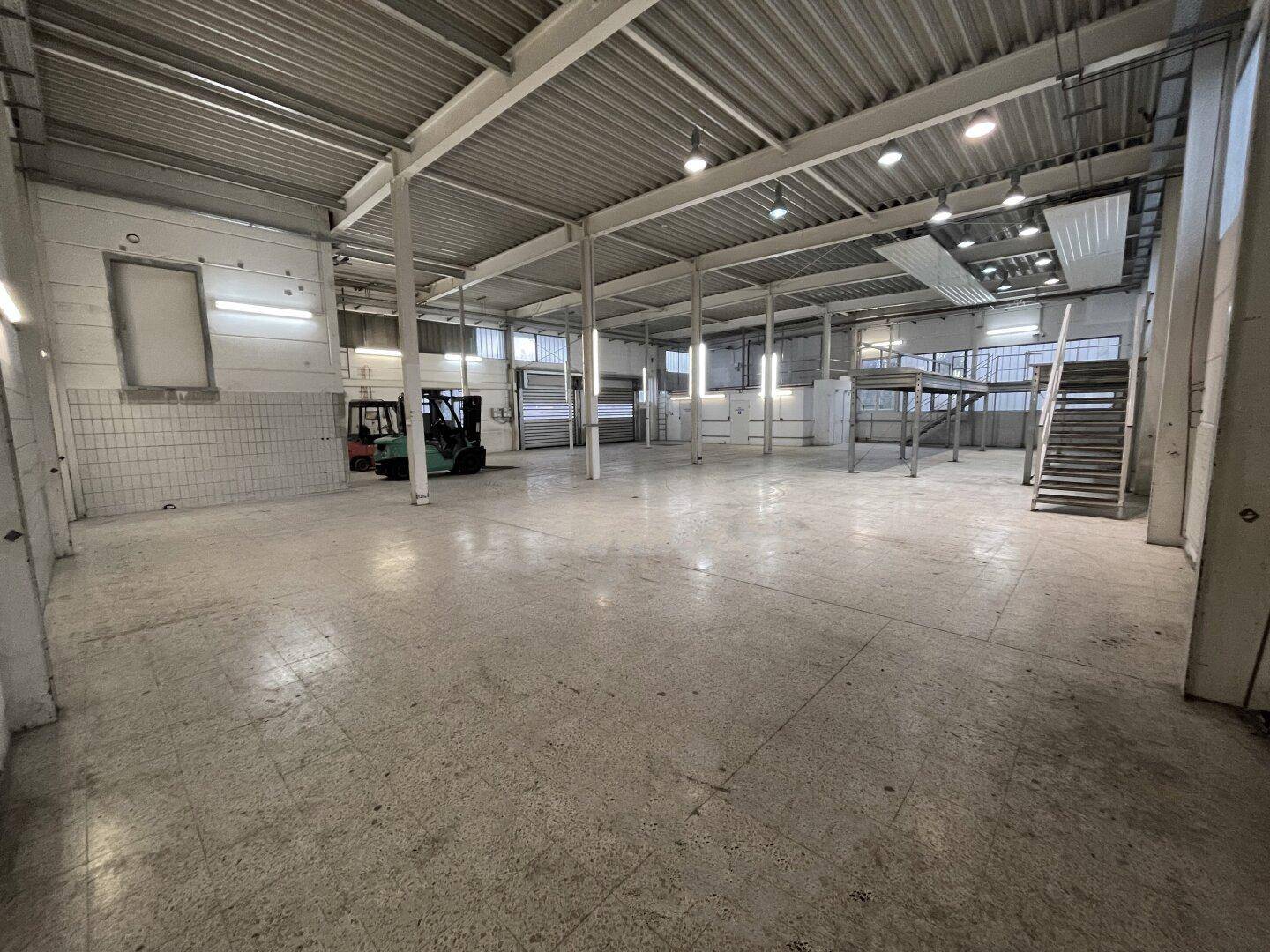 Große, leere Lagerhalle mit hohen Decken, Industrieboden und einer Metalltreppe zu einer Empore.