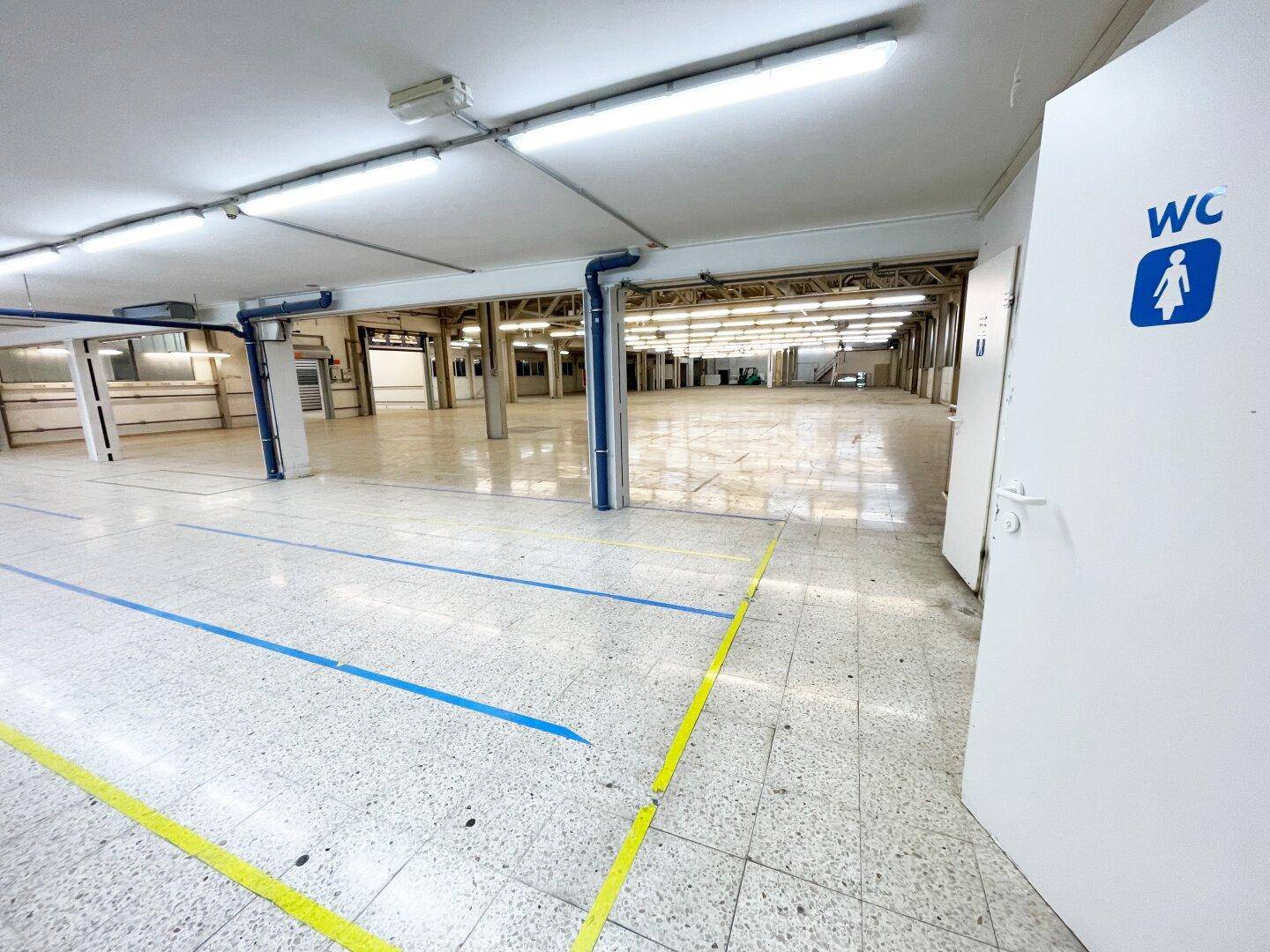Großer, sauberer Lagerbereich mit hohen Decken, Industrieboden und einer WC-Tür im Vordergrund.